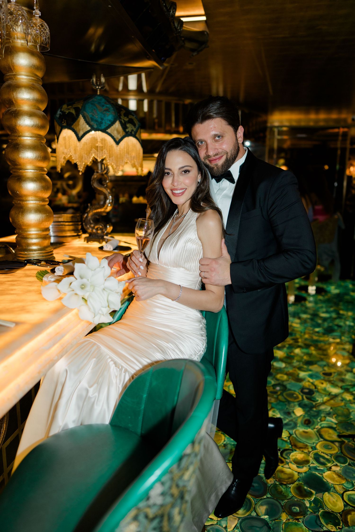 Elegant couple in wedding attire smiling at a luxurious bar, bride in white gown holding champagne, groom in black tuxedo.
