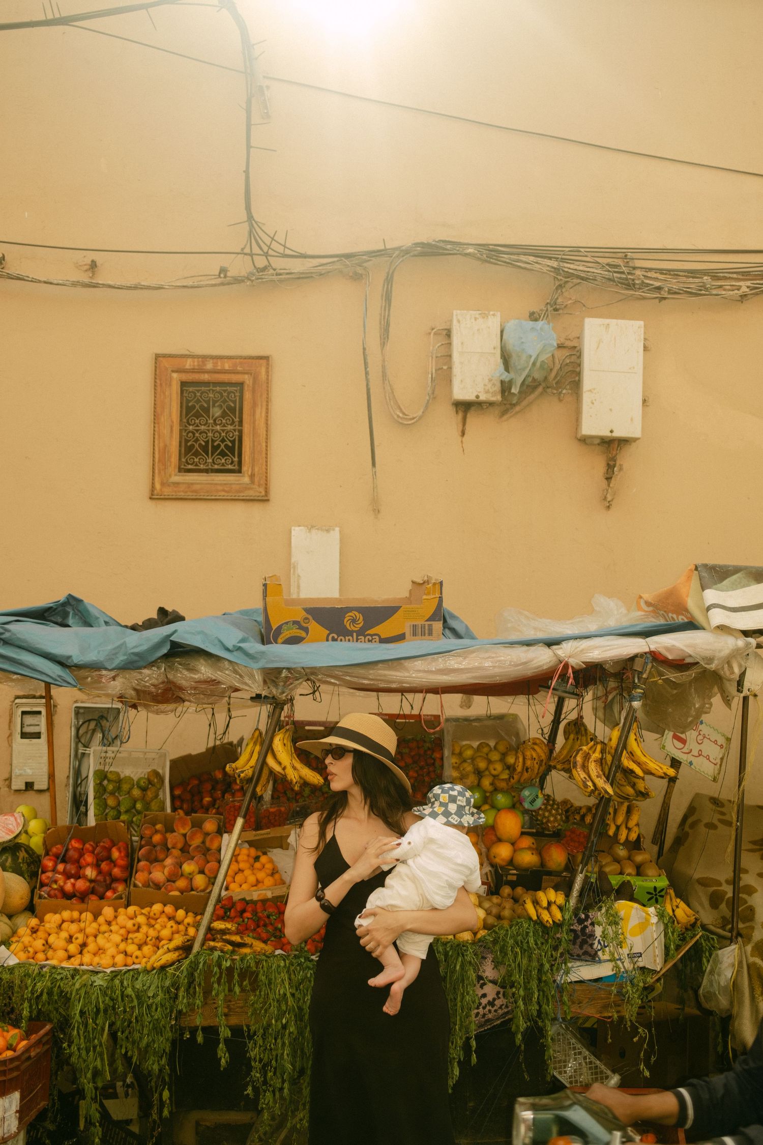 Woman in sun hat holding baby at colorful Moroccan fruit market stall with fresh produce display.