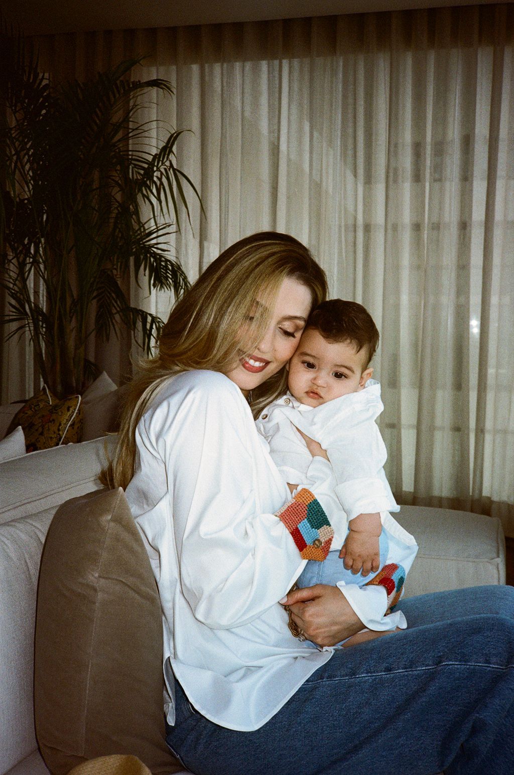 A smiling woman in a white shirt holds a baby dressed in white on a cozy sofa near sheer curtains and a palm plant.