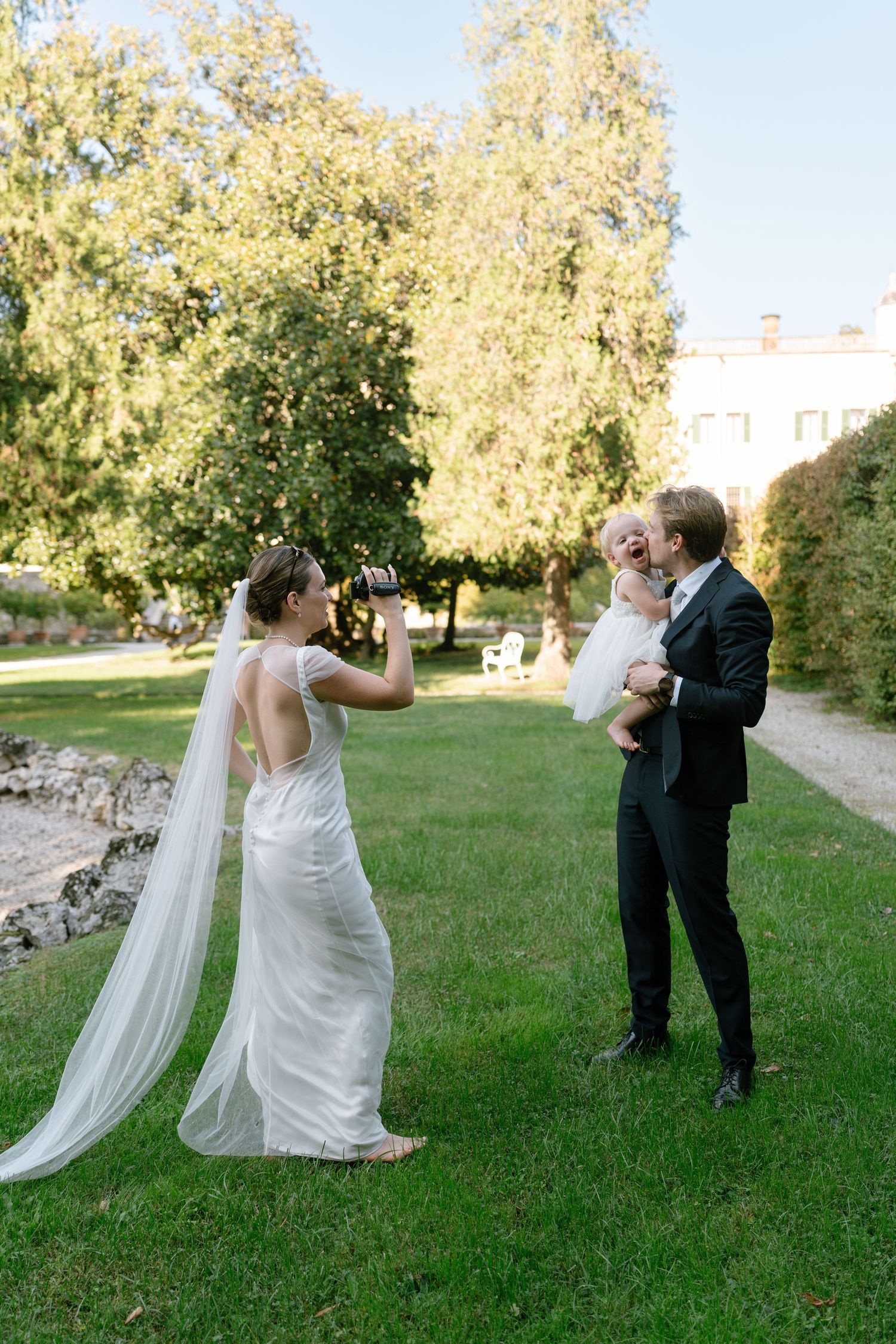 A wedding photographer captures a candid moment on a lawn with guests and a flowing white dress.