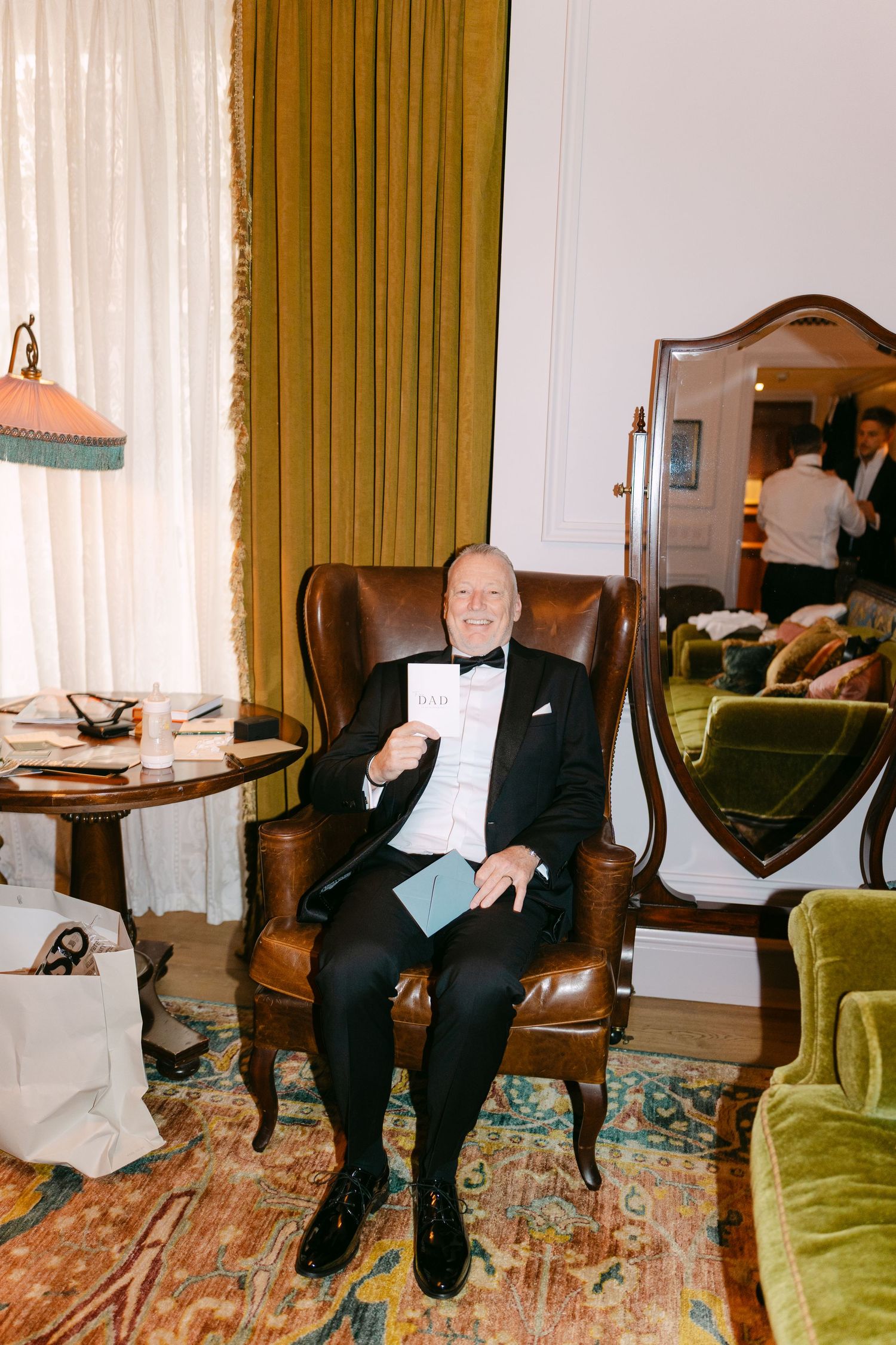 A person sits in a leather armchair holding papers, dressed in formal evening attire.