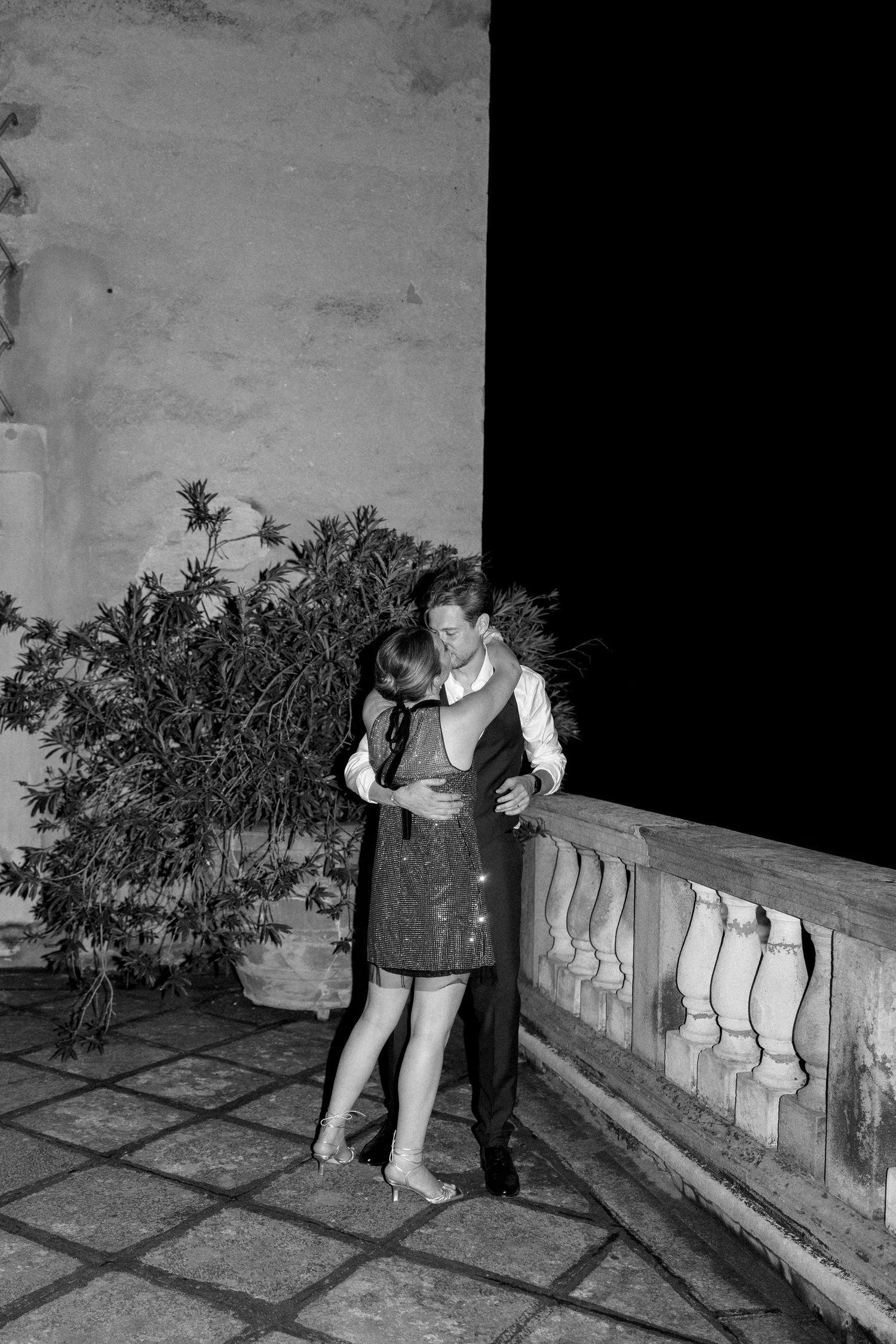 A couple embraces romantically against a stone wall in a vintage black and white photograph.