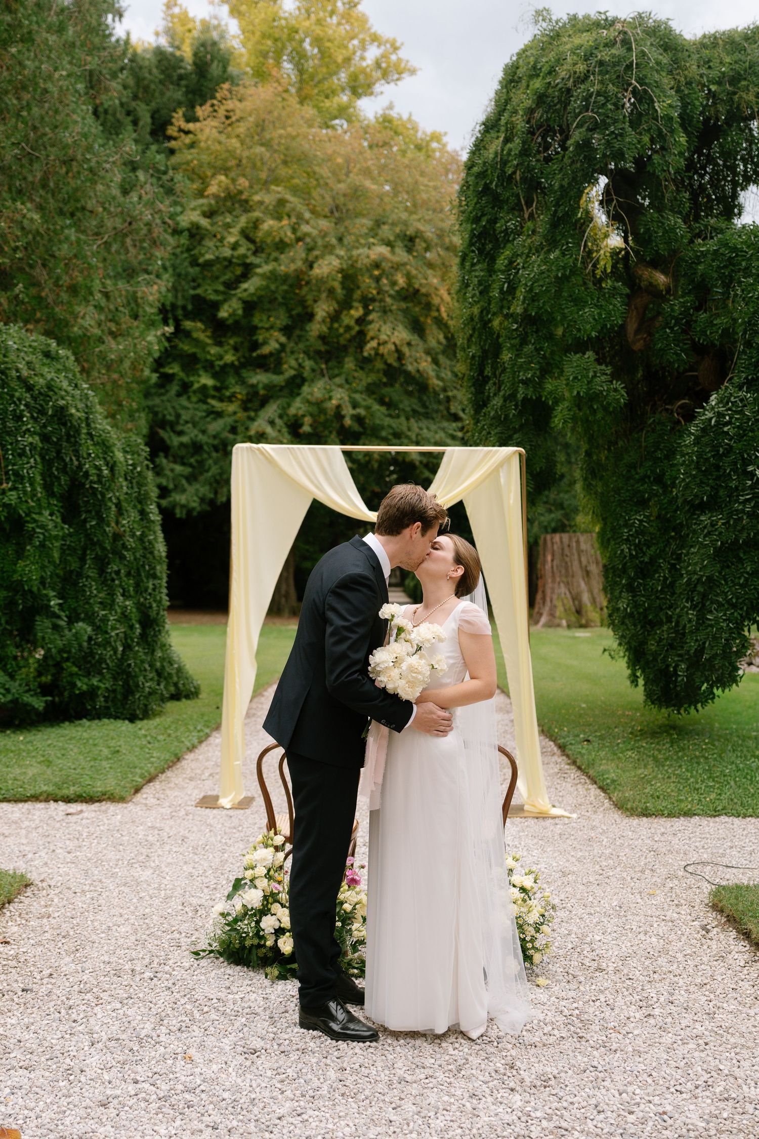 Newlyweds share a kiss under an elegant cream-colored arch on a gravel pathway surrounded by lush gardens.