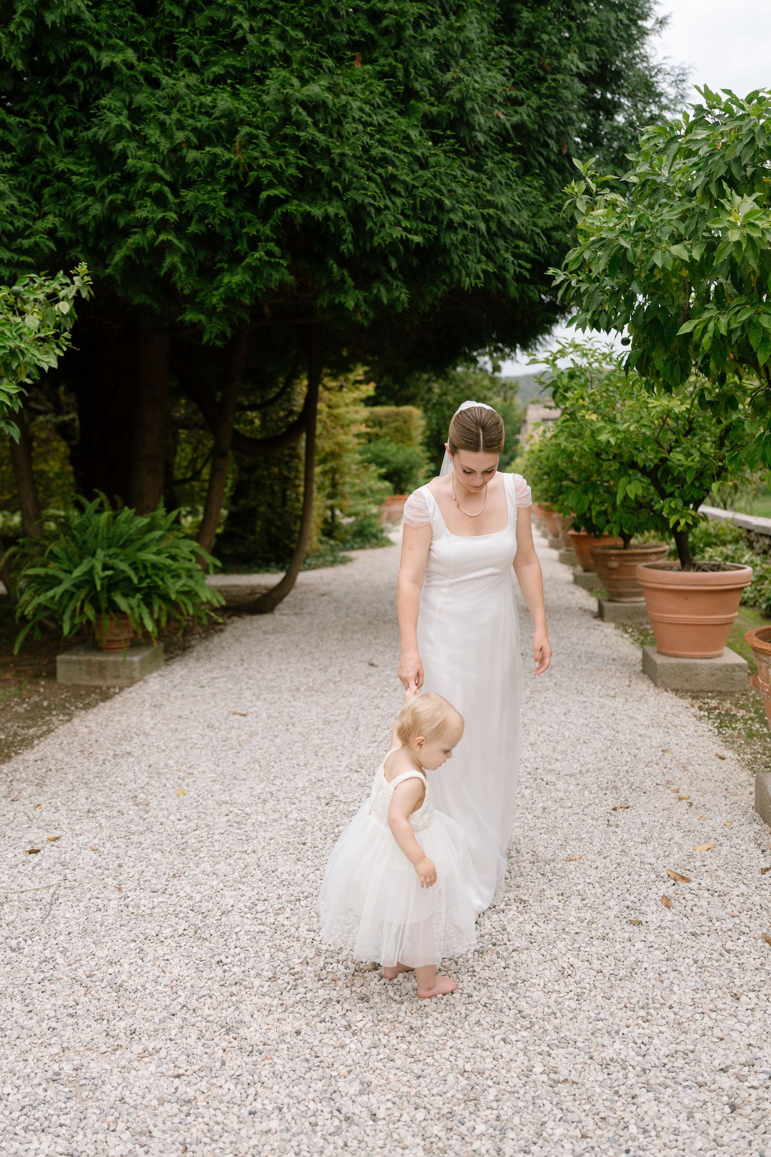 A person in a white dress walks with a small child along a gravel garden path.