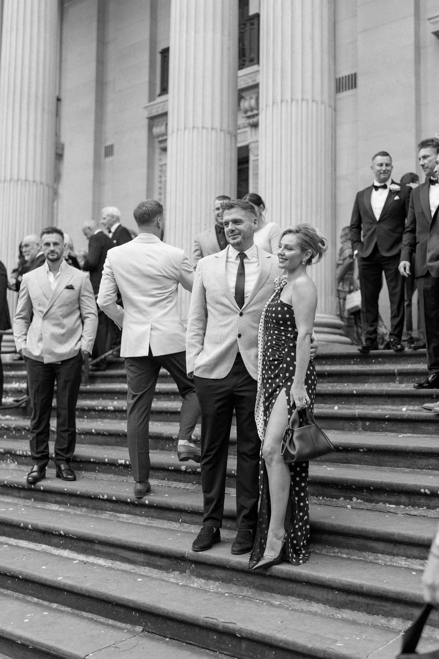 Elegantly dressed guests gather on grand stone steps with classical columns in black and white photography.