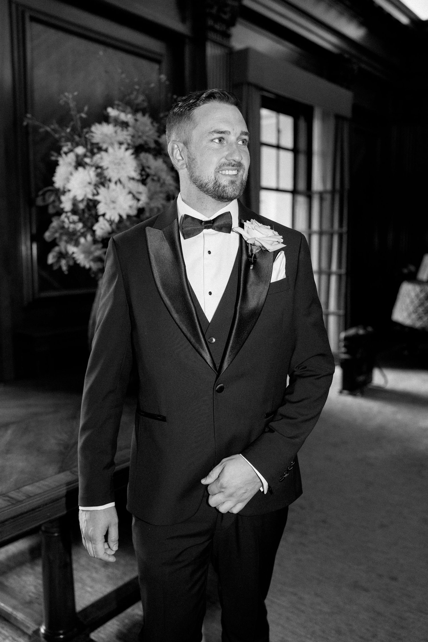 Groomsmen in formal black tuxedos and bowties stand in a wood-paneled hallway.
