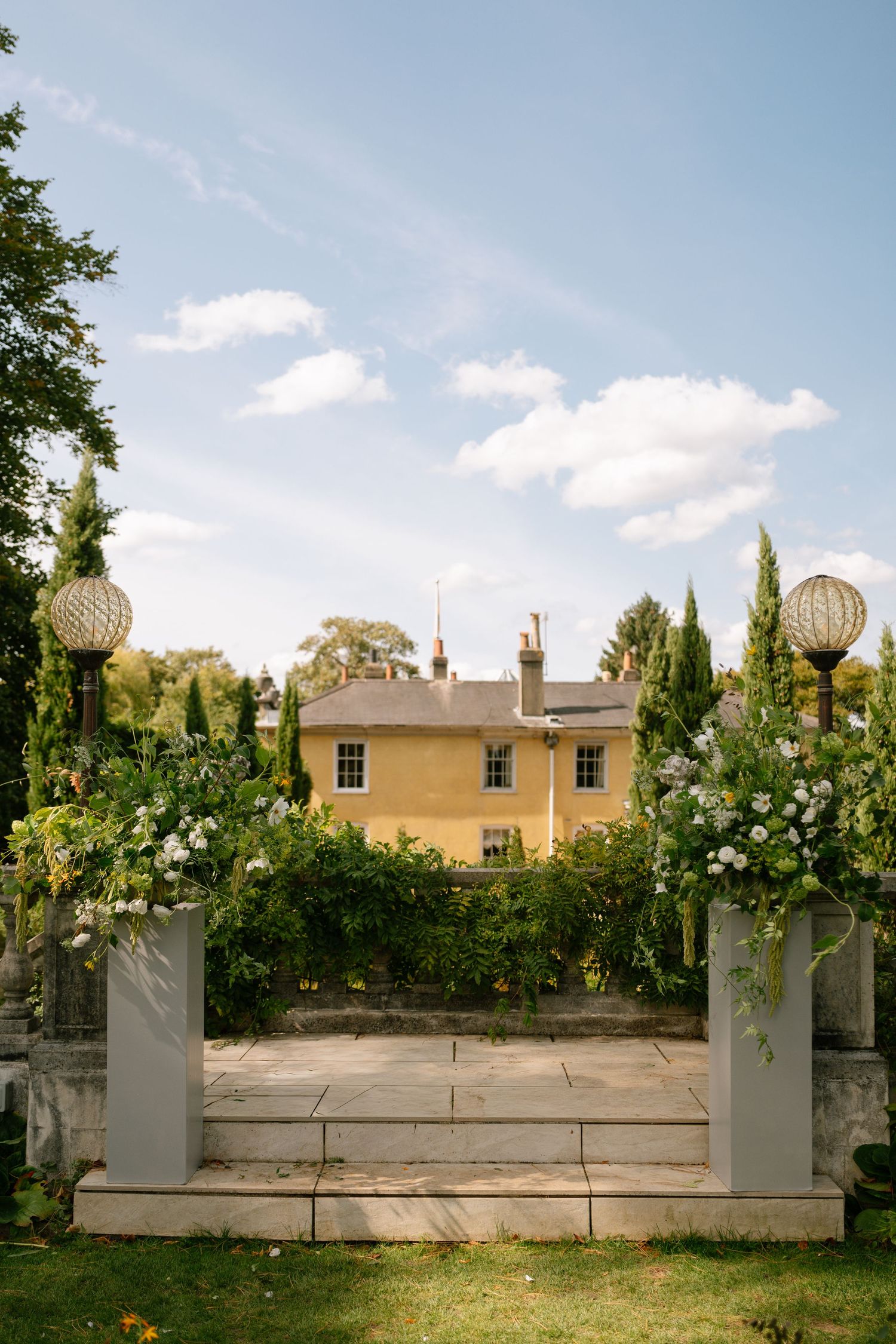 Wedding ceremony space with stone pillars, floral arrangements, and manor house backdrop.