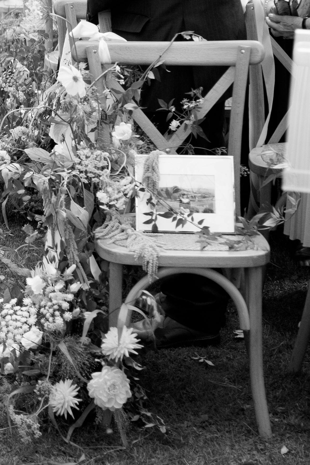 Black and white detail of floral arrangements decorating wooden chairs at an outdoor wedding.