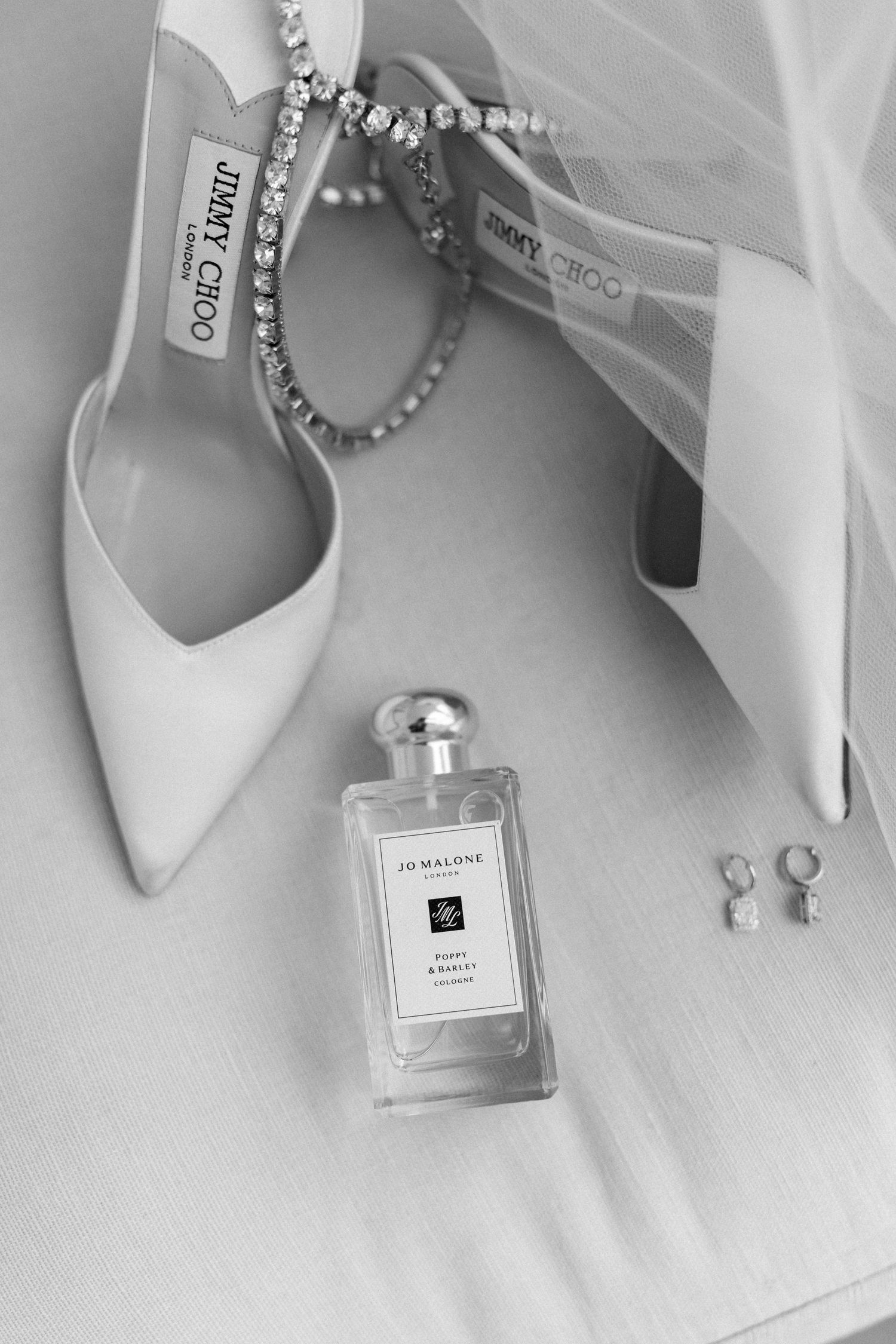 Wedding accessories including perfume bottles, jewelry and white shoes arranged on white fabric.