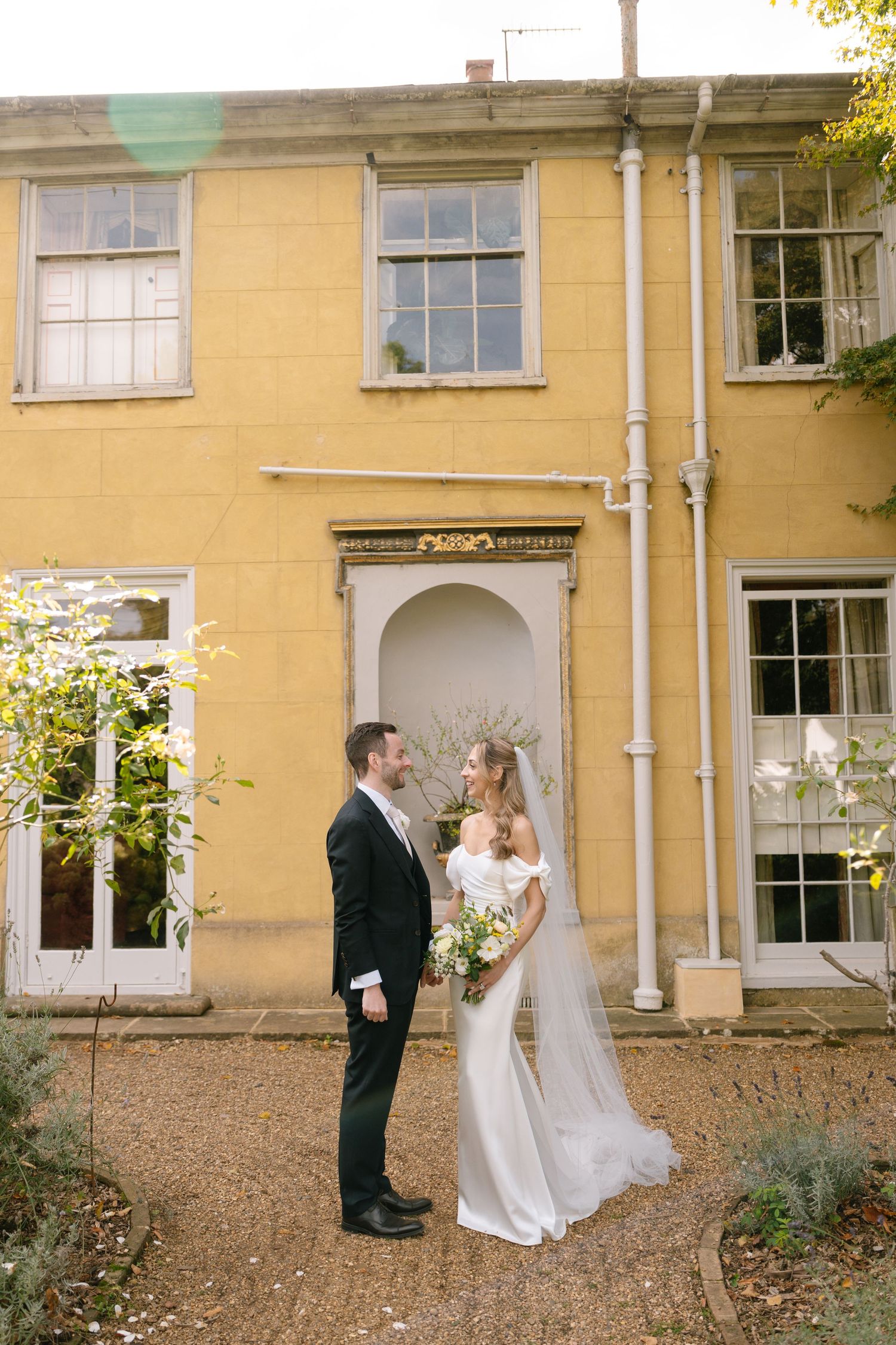 Couple in wedding attire stands outside historic yellow building with white trim windows.
