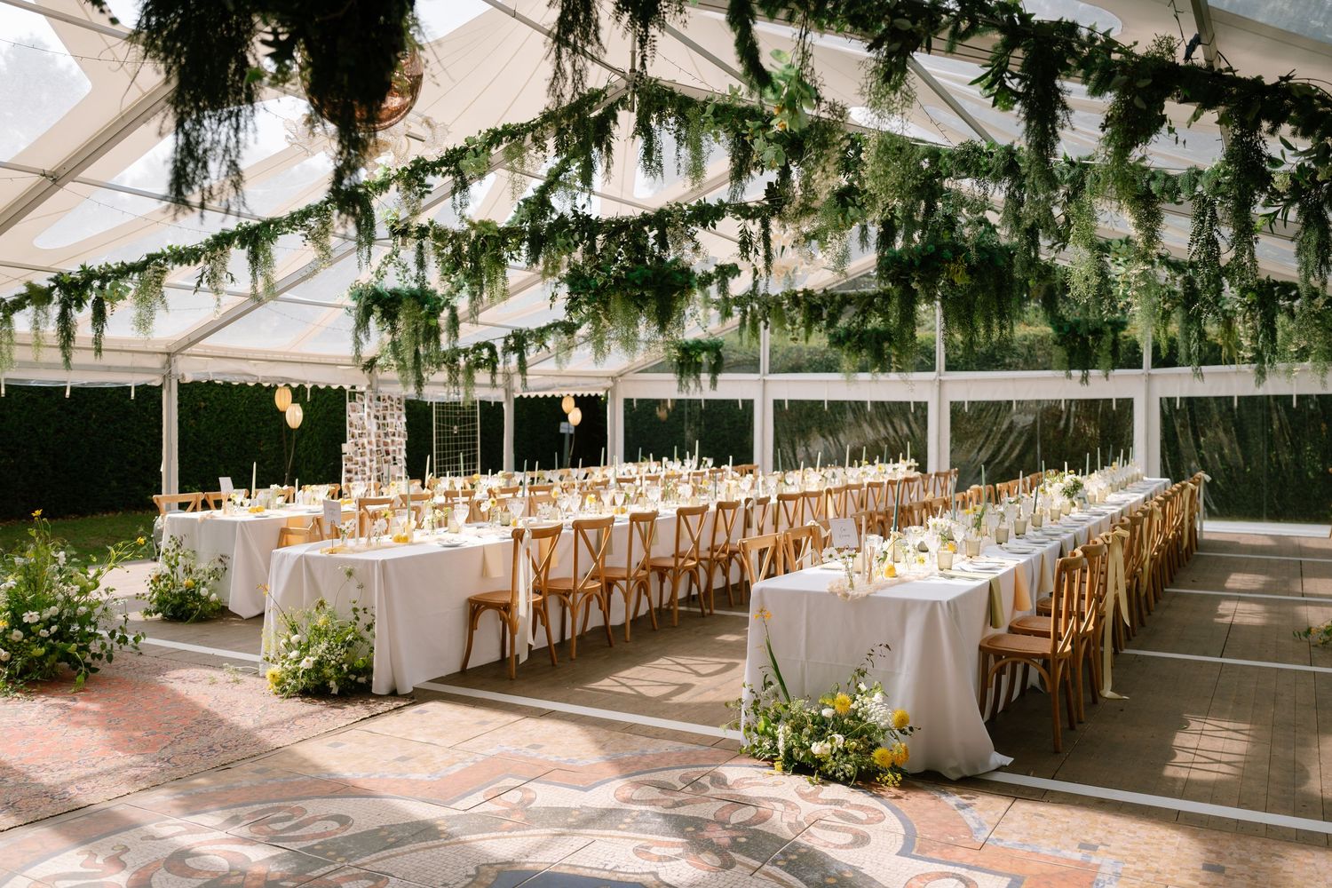 Long banquet tables set up under an elegant white tent canopy with greenery hanging from above.