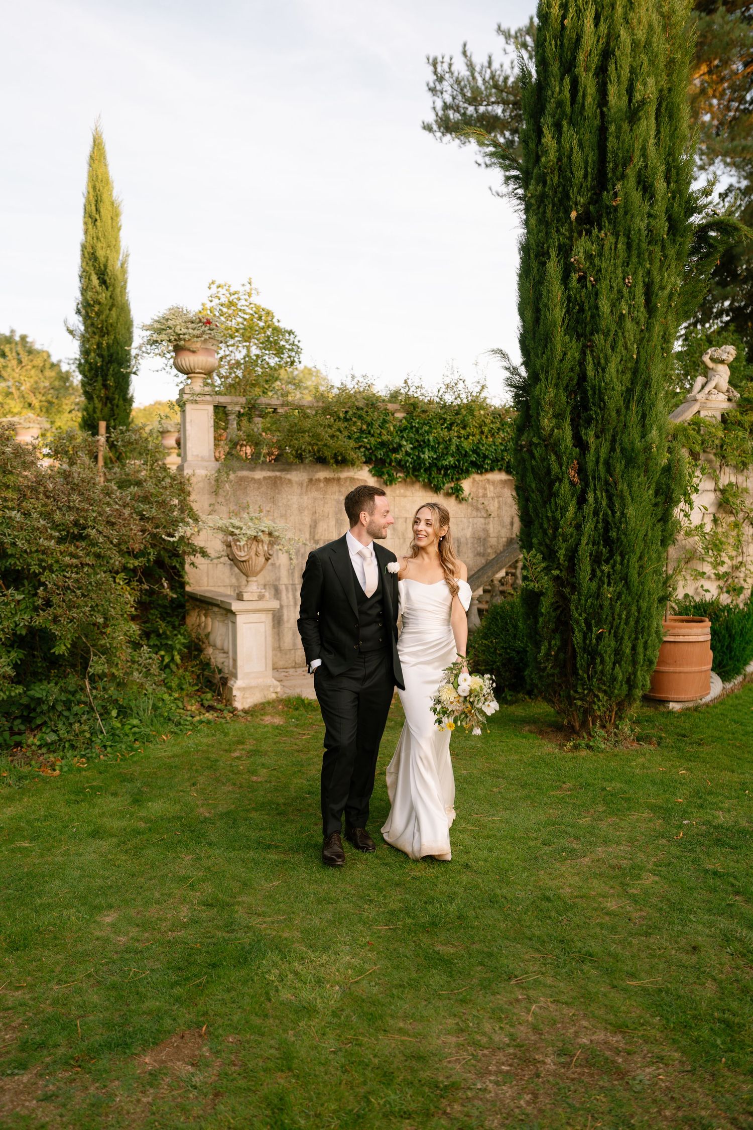 Couple poses in garden with tall cypress trees and stone wall in background.