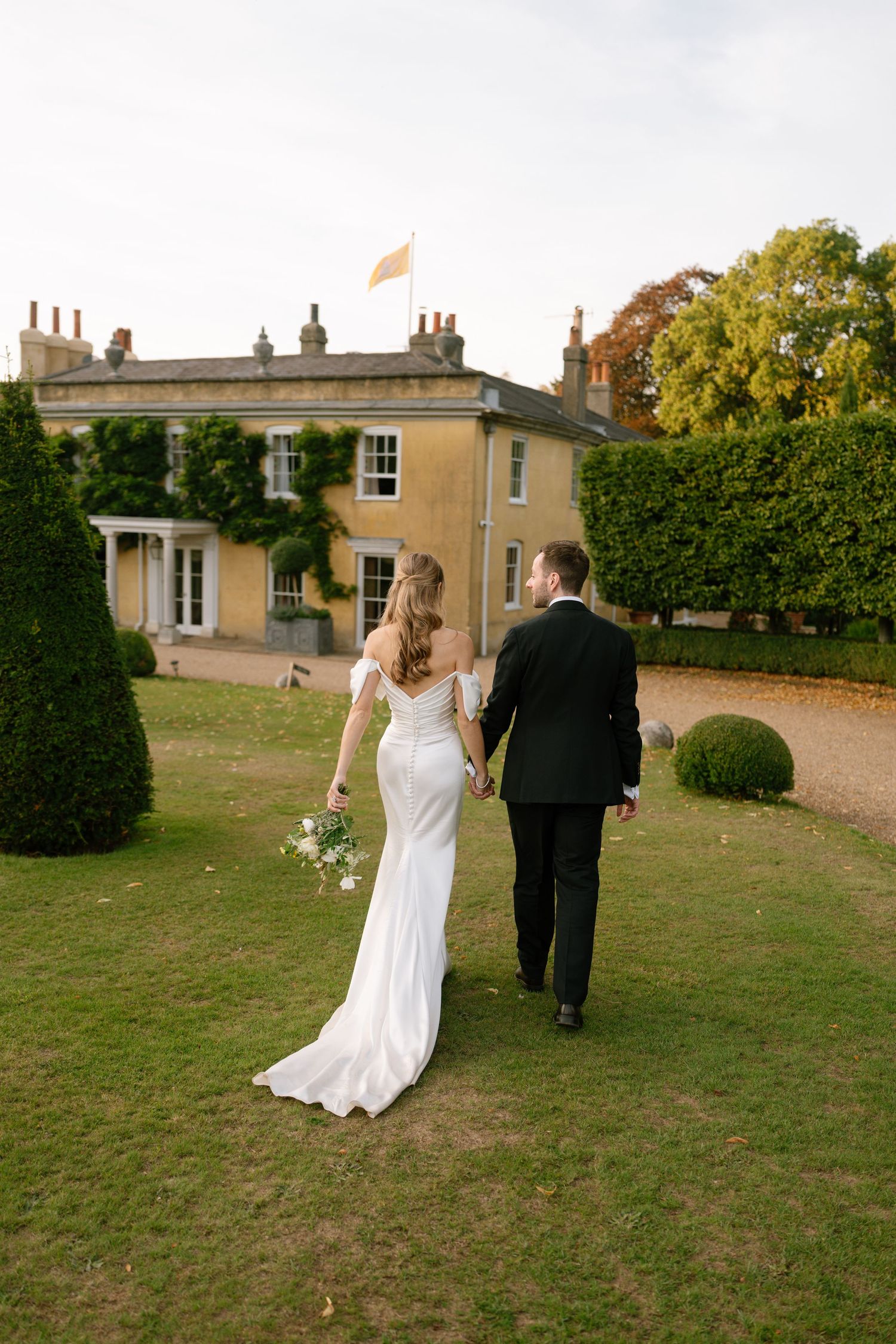 A wedding couple walks across a manicured lawn towards a grand country house.