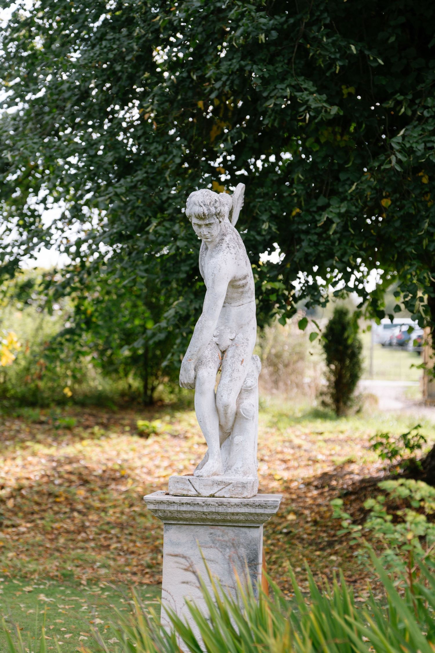 Classical white garden statue nestled among trees and fallen leaves.