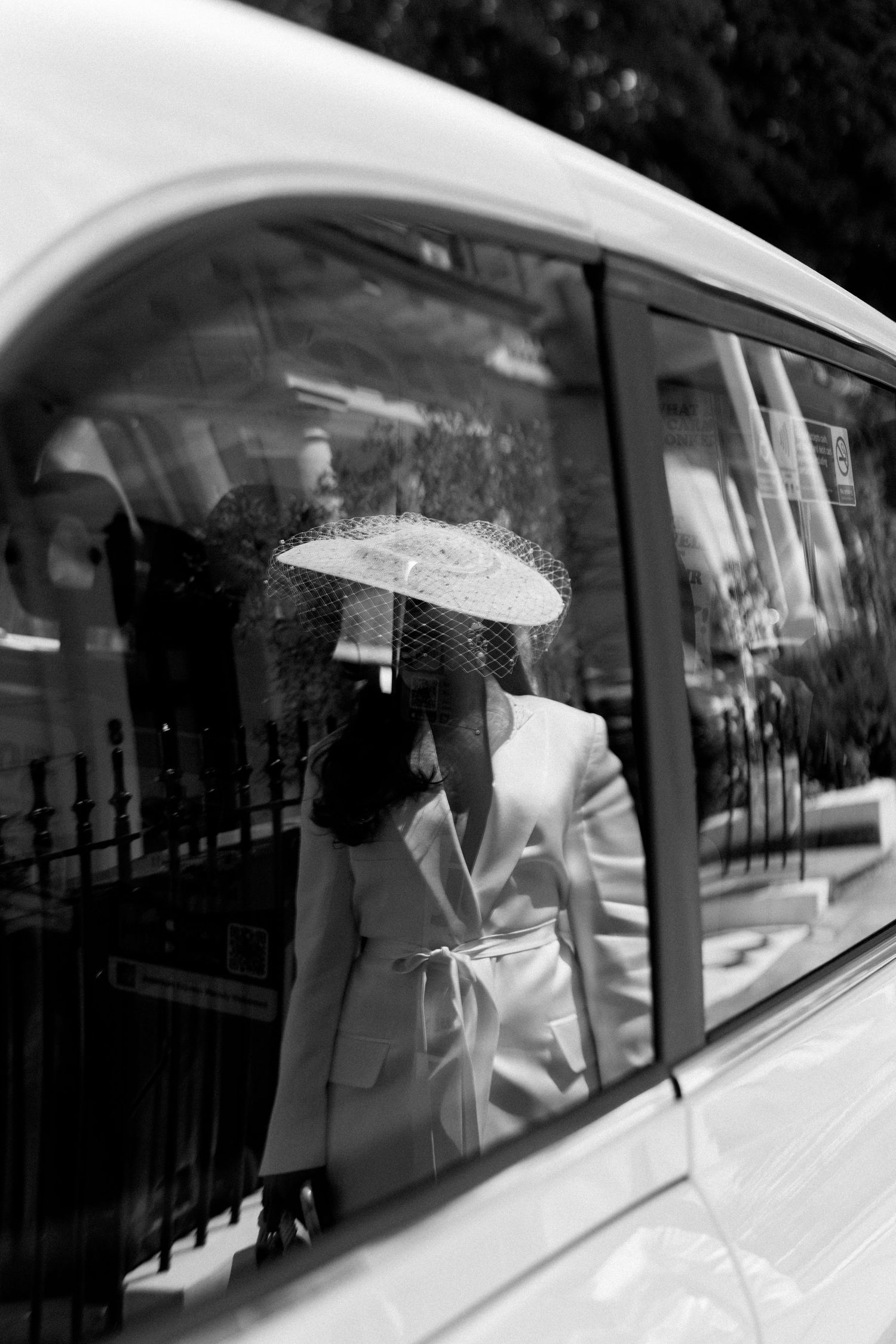 Black and white photo of an elegant bride in white suit and veiled hat seen through a car window.