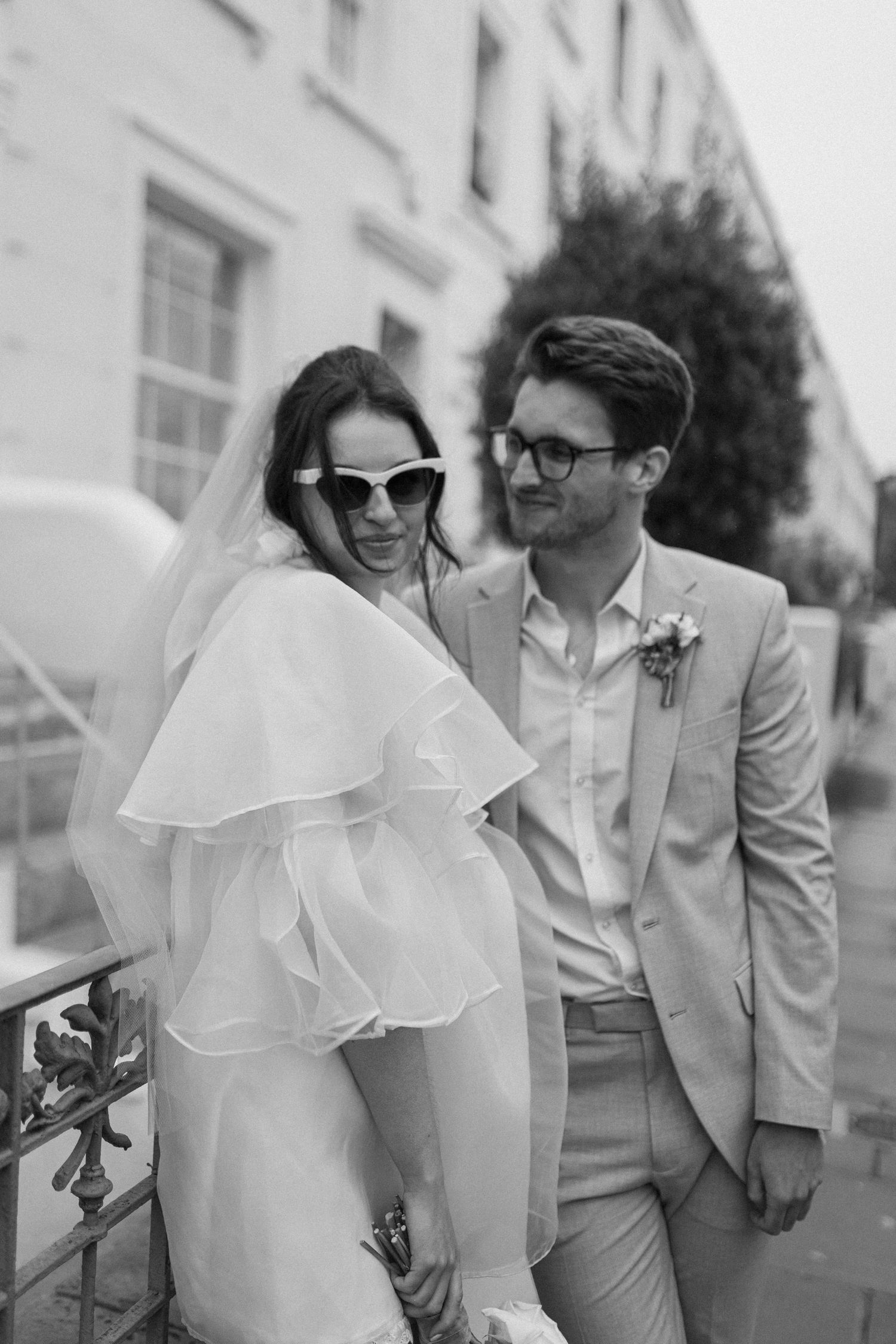 Black and white photo of a stylish bride in sunglasses and groom in suit smiling together on a city street.
