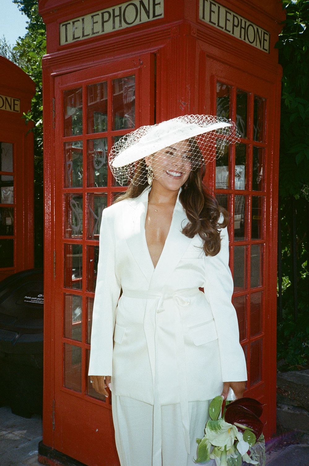 Woman in white suit and wide-brim hat with netting poses smiling in front of red British telephone box.
