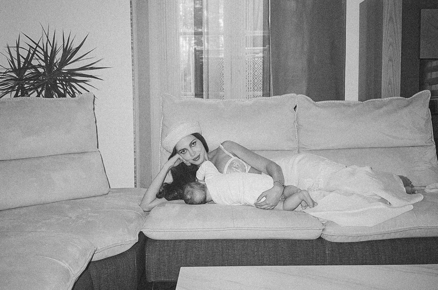 Black and white photo of a woman in white dress and hat relaxing on a beige sectional sofa indoors.