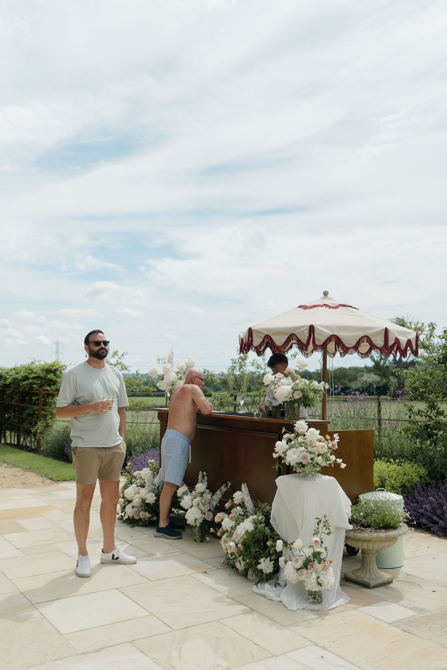 Outdoor wedding bar cart decorated with white florals under striped umbrella at English countryside venue.