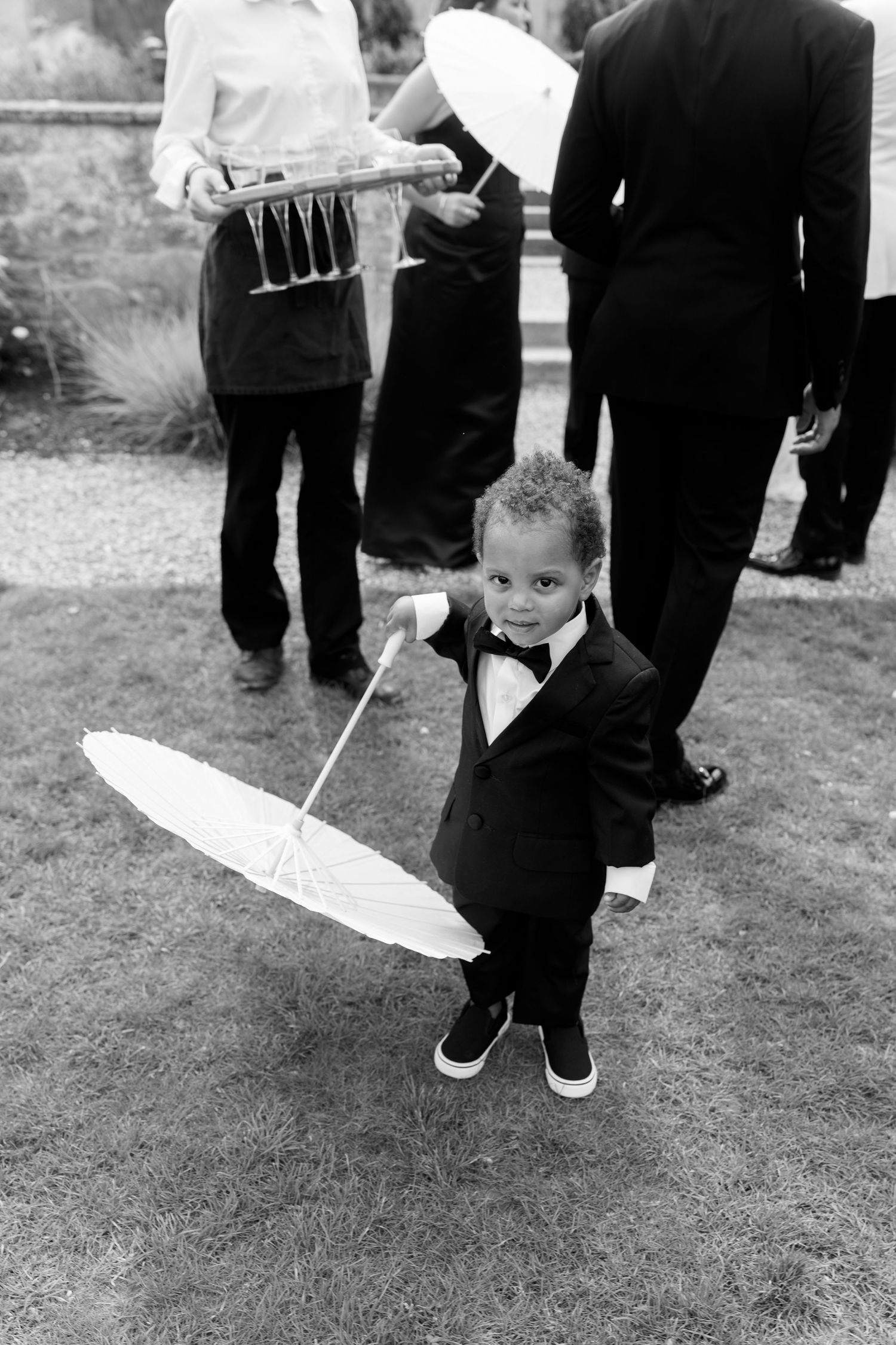 An adorable young boy in a tuxedo holds a white parasol umbrella at a wedding reception.