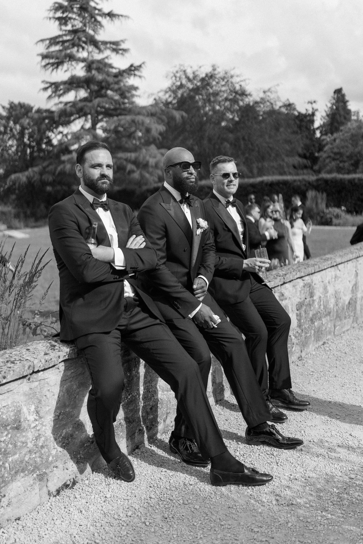 Three groomsmen in black tuxedos with bow ties sit on stone wall at outdoor wedding in black and white.