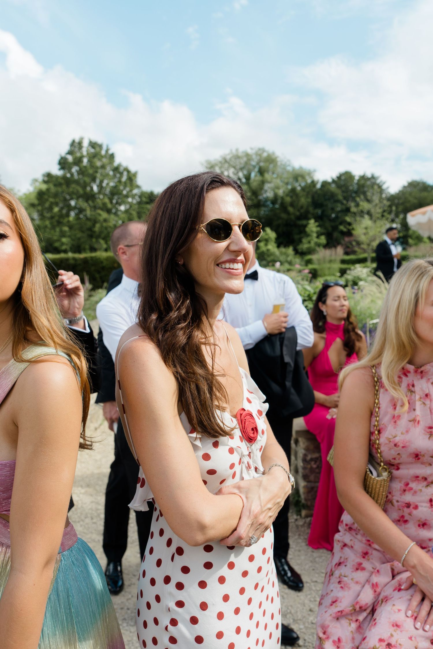 Smiling brunette woman in a polka dot dress wearing sunglasses at an outdoor summer wedding.