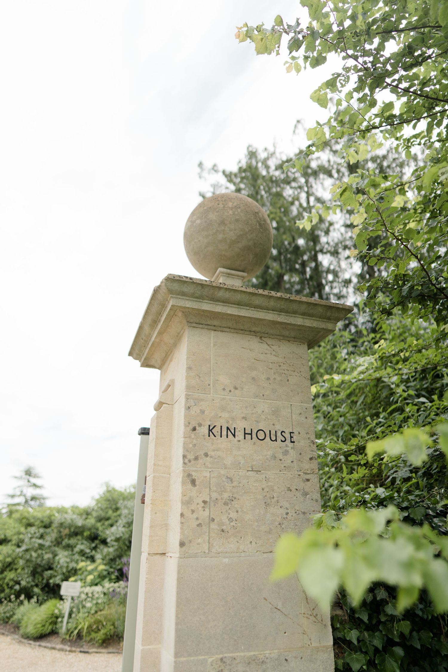 Stone gate pillar with spherical finial engraved with Kin House surrounded by lush green trees.