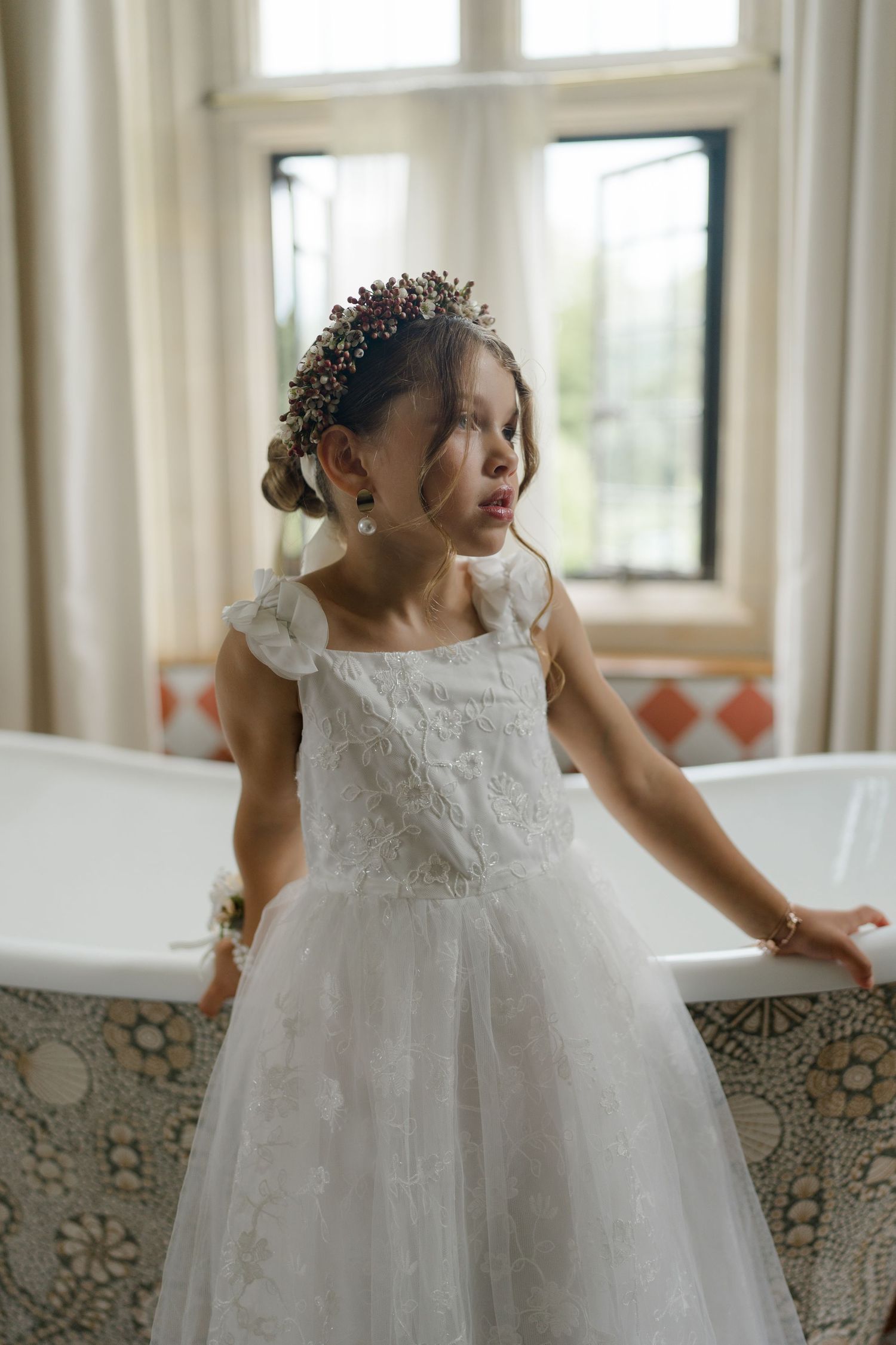 Young flower girl in white lace dress with floral crown standing beside a decorative freestanding bathtub.