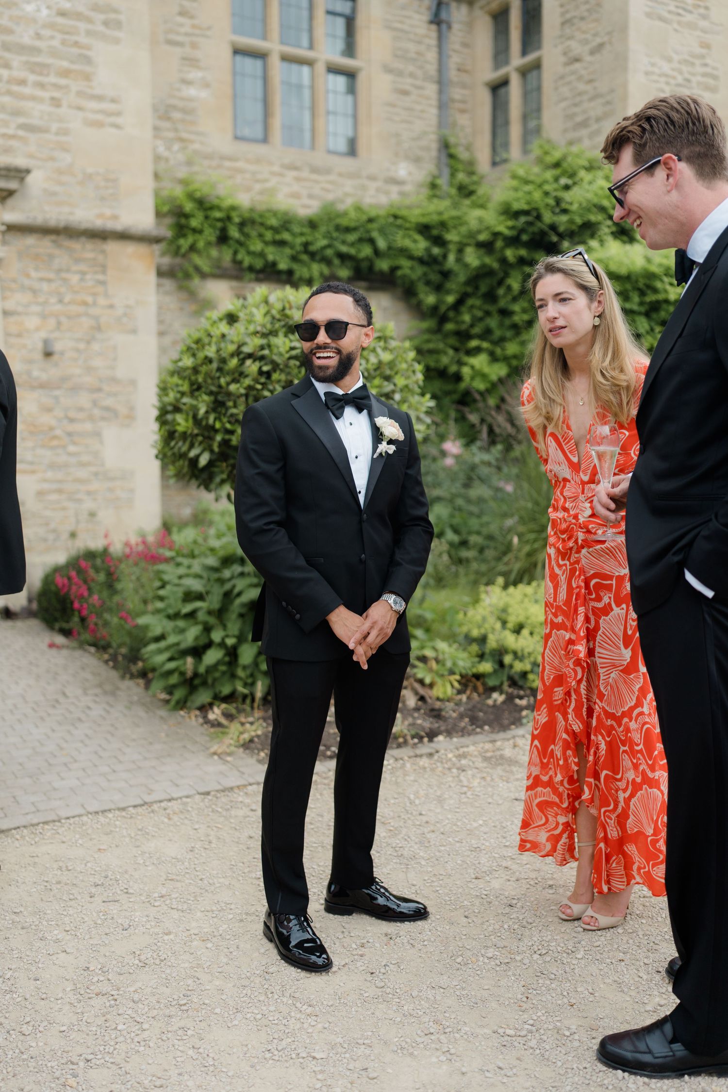 Groom in a black tuxedo with sunglasses smiling while greeting guests outside a manor.