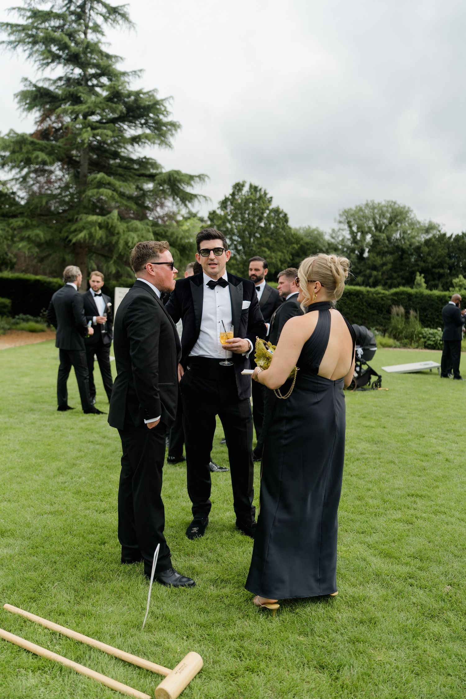 Wedding guests in black tie attire socializing on lawn with croquet mallets during outdoor reception.