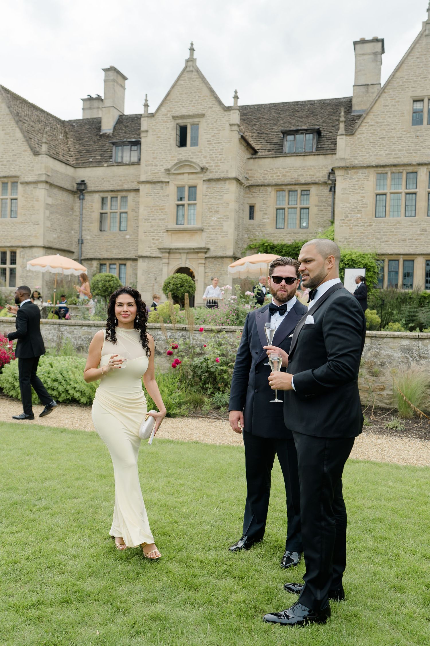 Elegant wedding guests in formal attire socializing on manicured lawn outside a grand stone manor house.