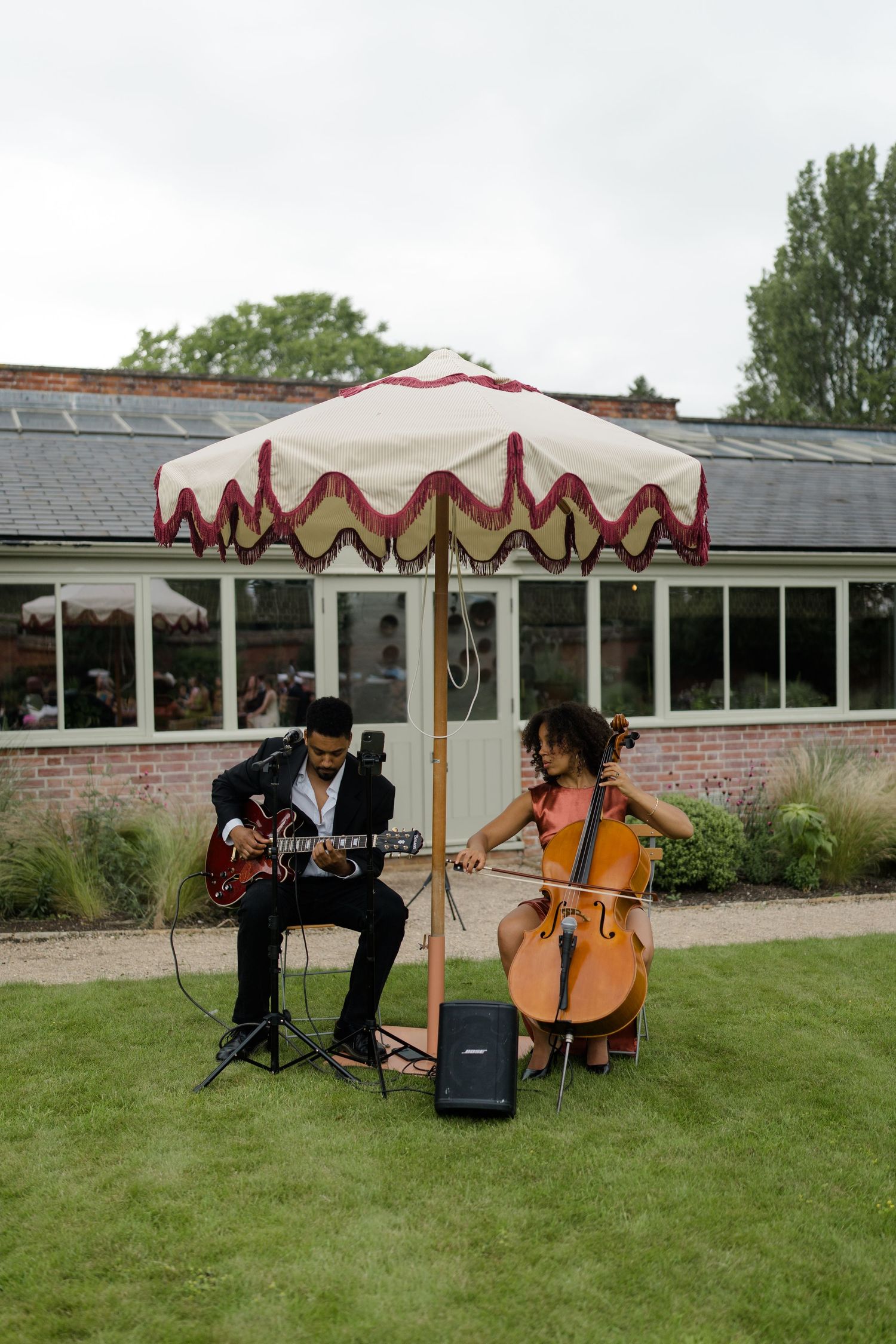 Two musicians perform outdoors at a wedding, one playing guitar and one playing cello under a decorative parasol.