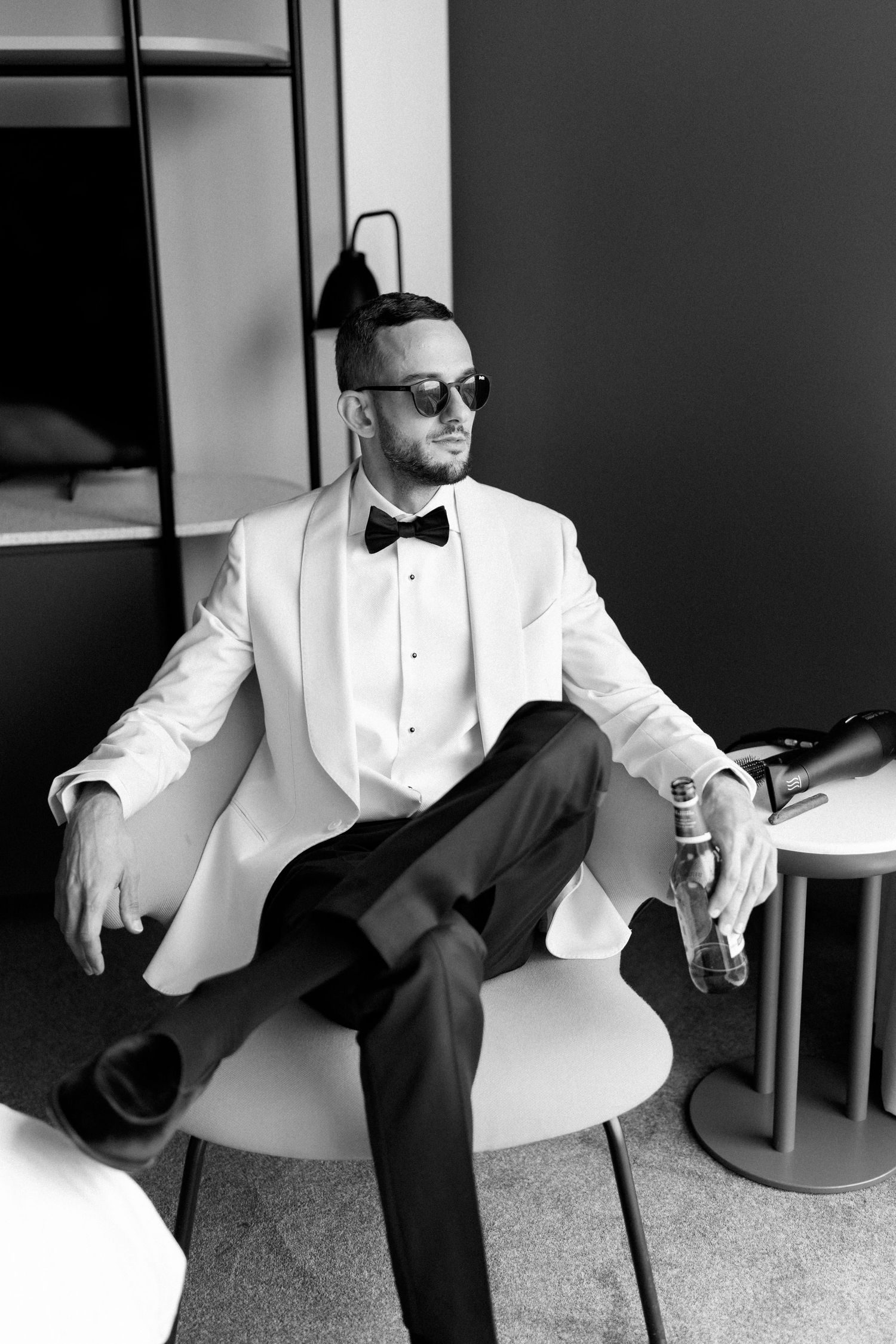Groom in white tuxedo jacket and bow tie poses in modern chair.