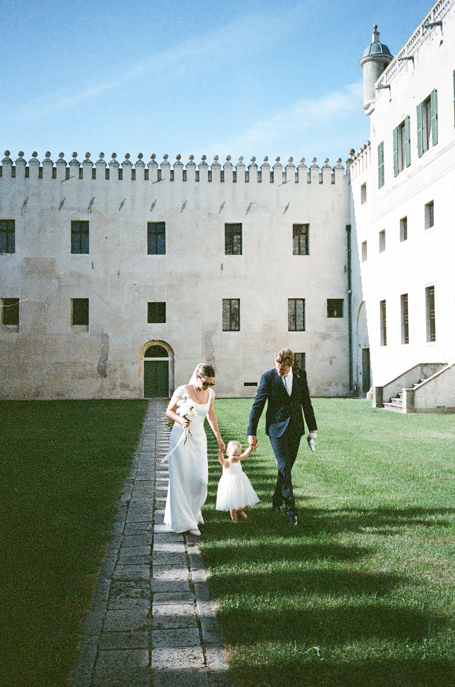 Three people walk along a stone path beside a white castle wall with green lawn on a sunny day.