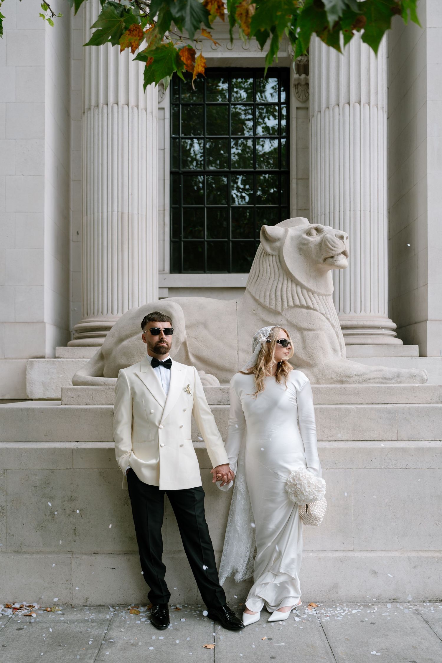 Couple in white formal wear poses playfully against a classical building with columns.