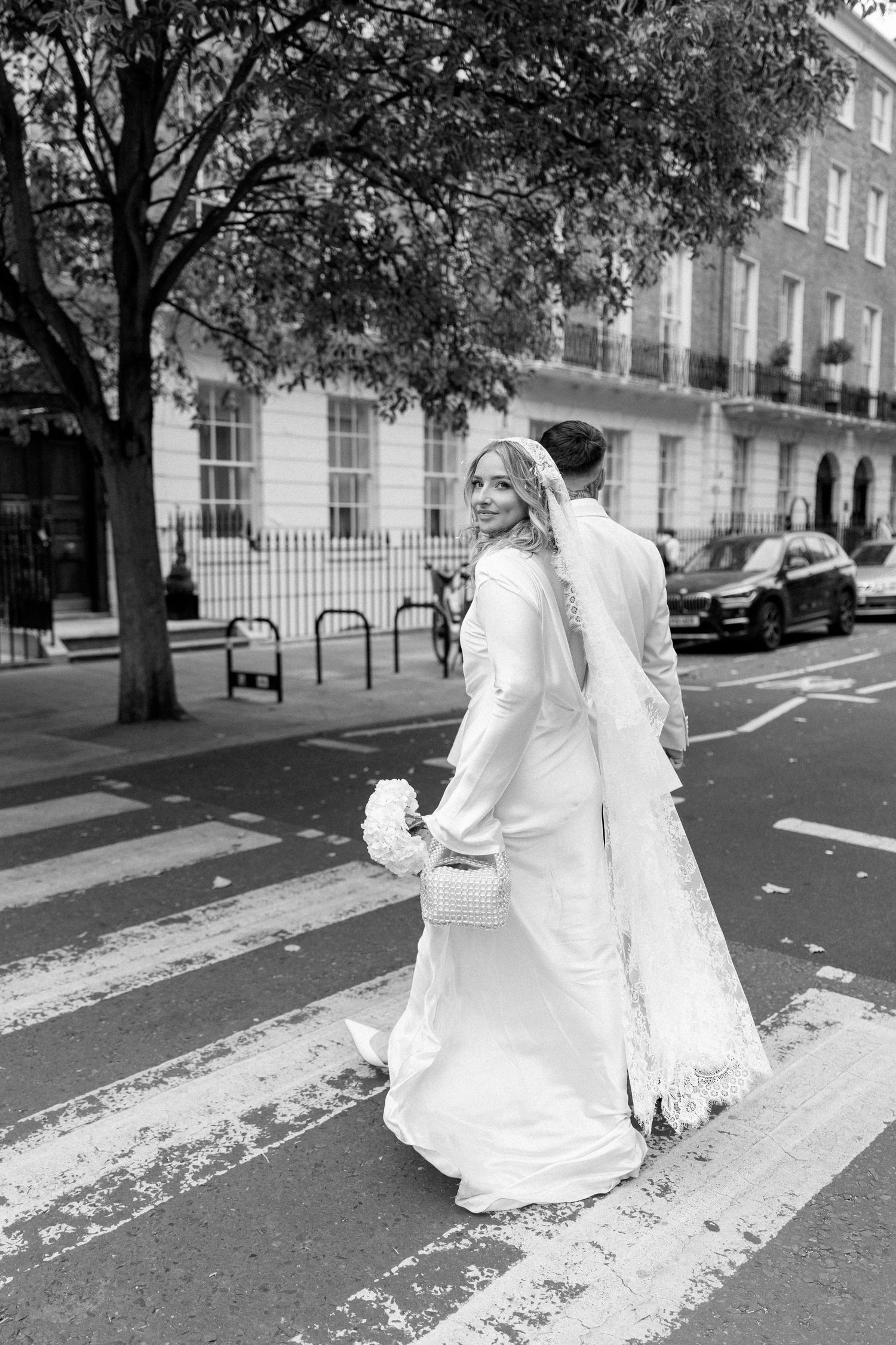 Bride in flowing white dress and veil crosses London street in black and white photograph.