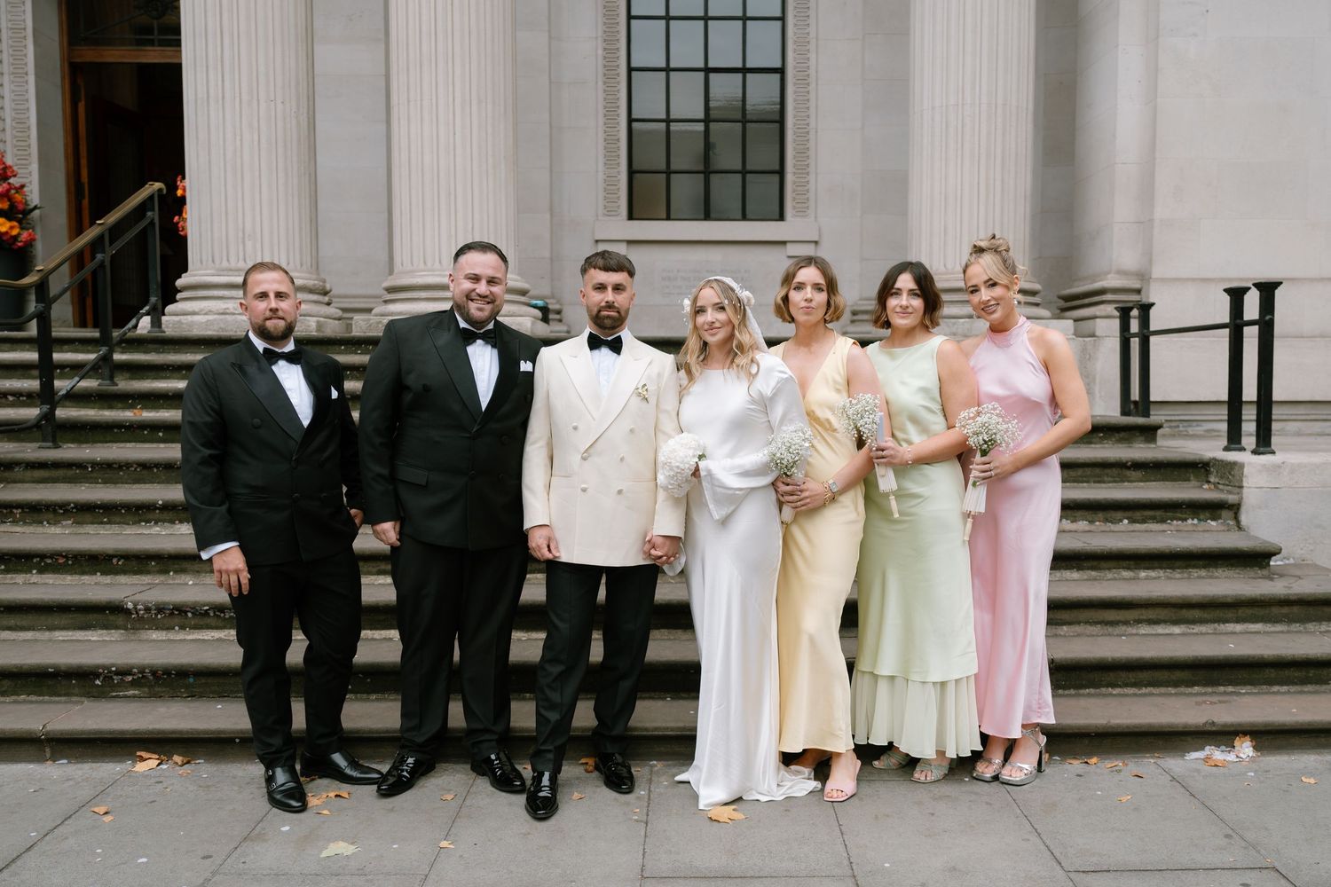 Wedding party poses on grand stone steps of a classical building with columns.