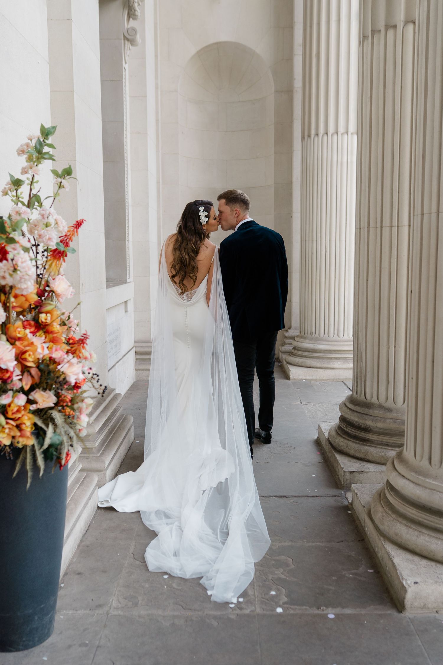 Wedding couple poses among classical columns with colorful floral arrangements.