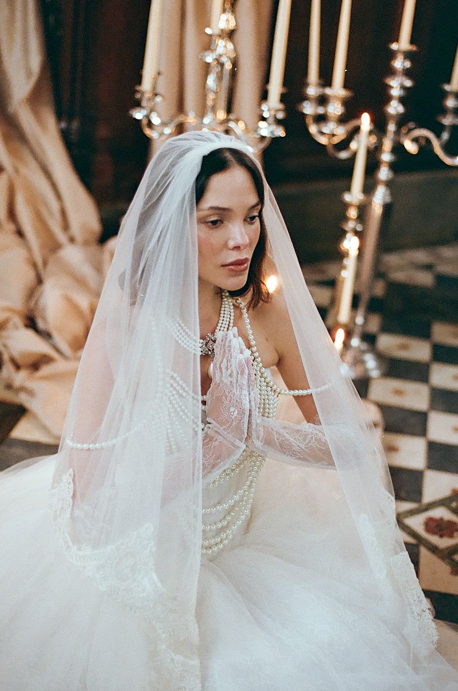 A bride in a white lace wedding dress and veil adorned with pearls poses elegantly in a classic setting.