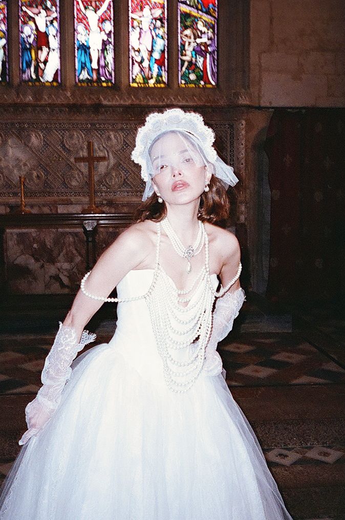 A figure in a white tulle gown and pearls poses dramatically in a gothic church interior with stained glass windows.