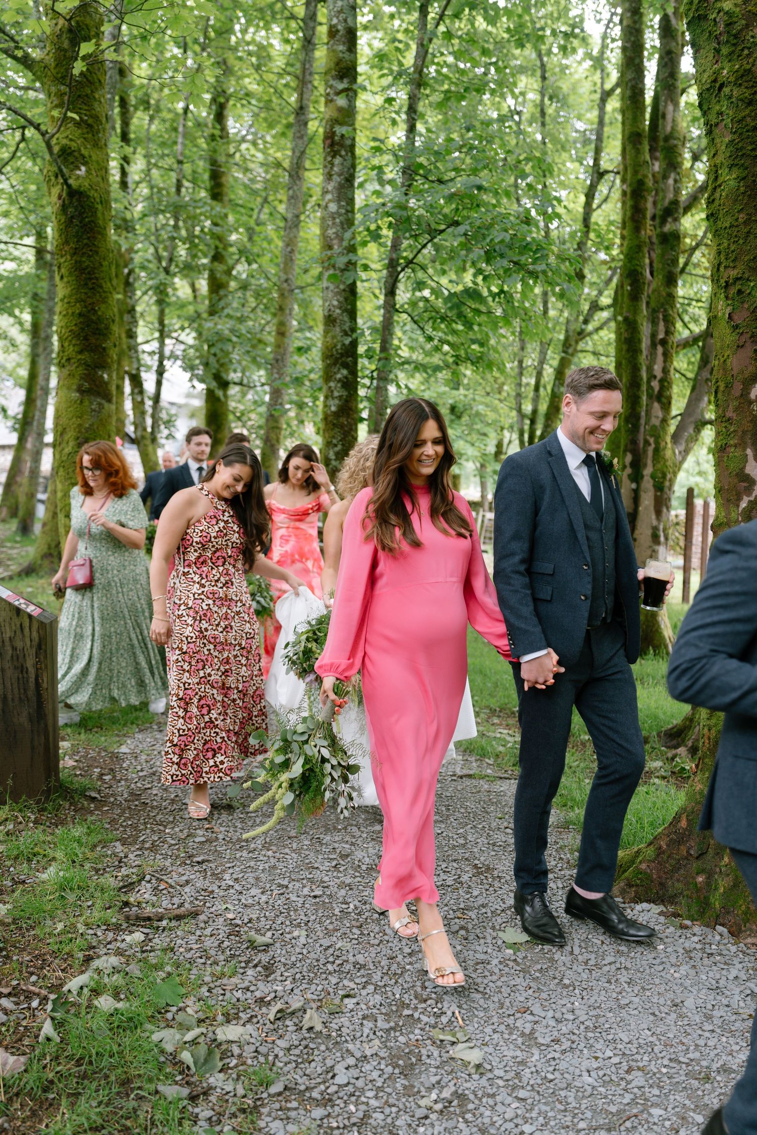 Wedding party walking along a forest path with guests in colorful dresses and formal attire.
