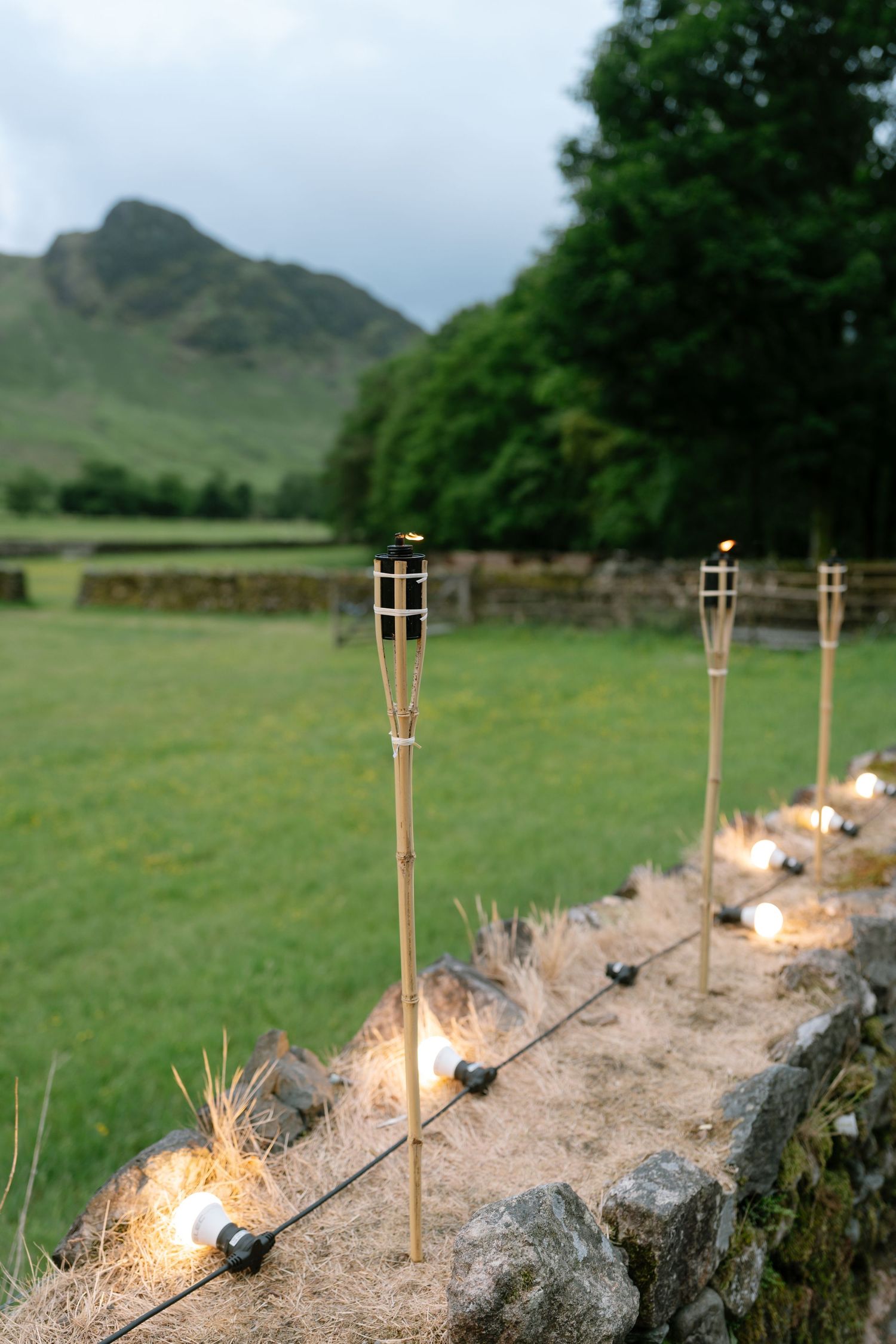 Lit tiki torches line a stone wall at dusk in a countryside setting.