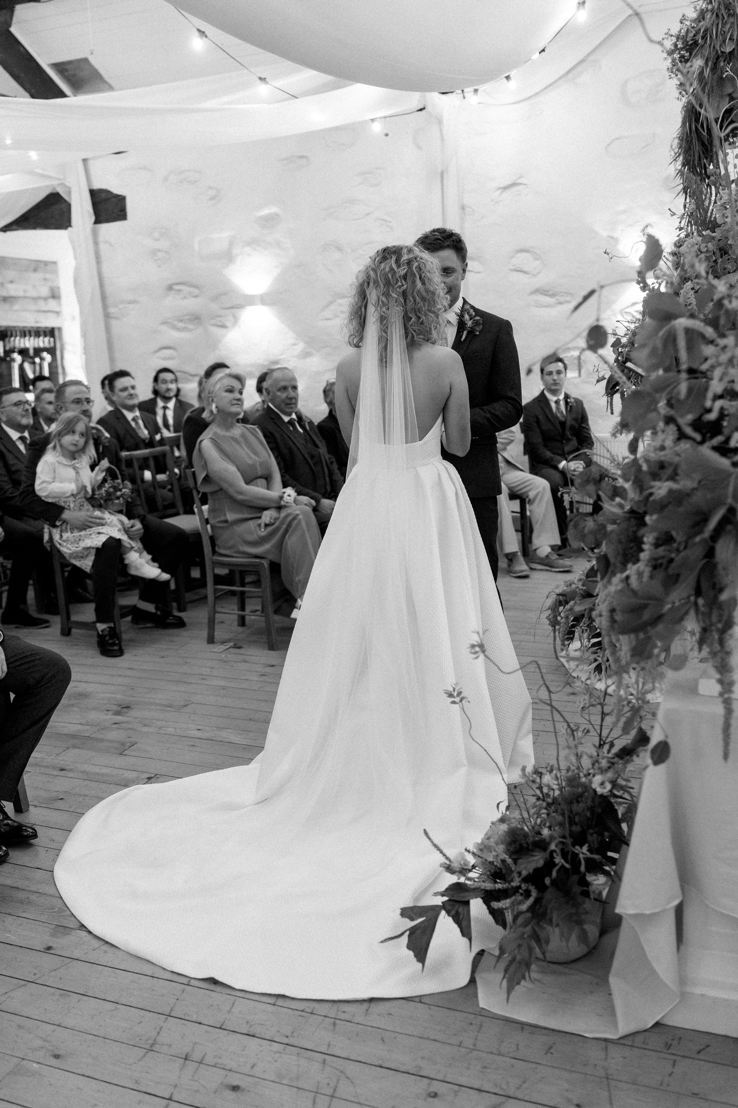 Wedding ceremony in a white-decorated venue with wooden floors and draped fabric ceiling.
