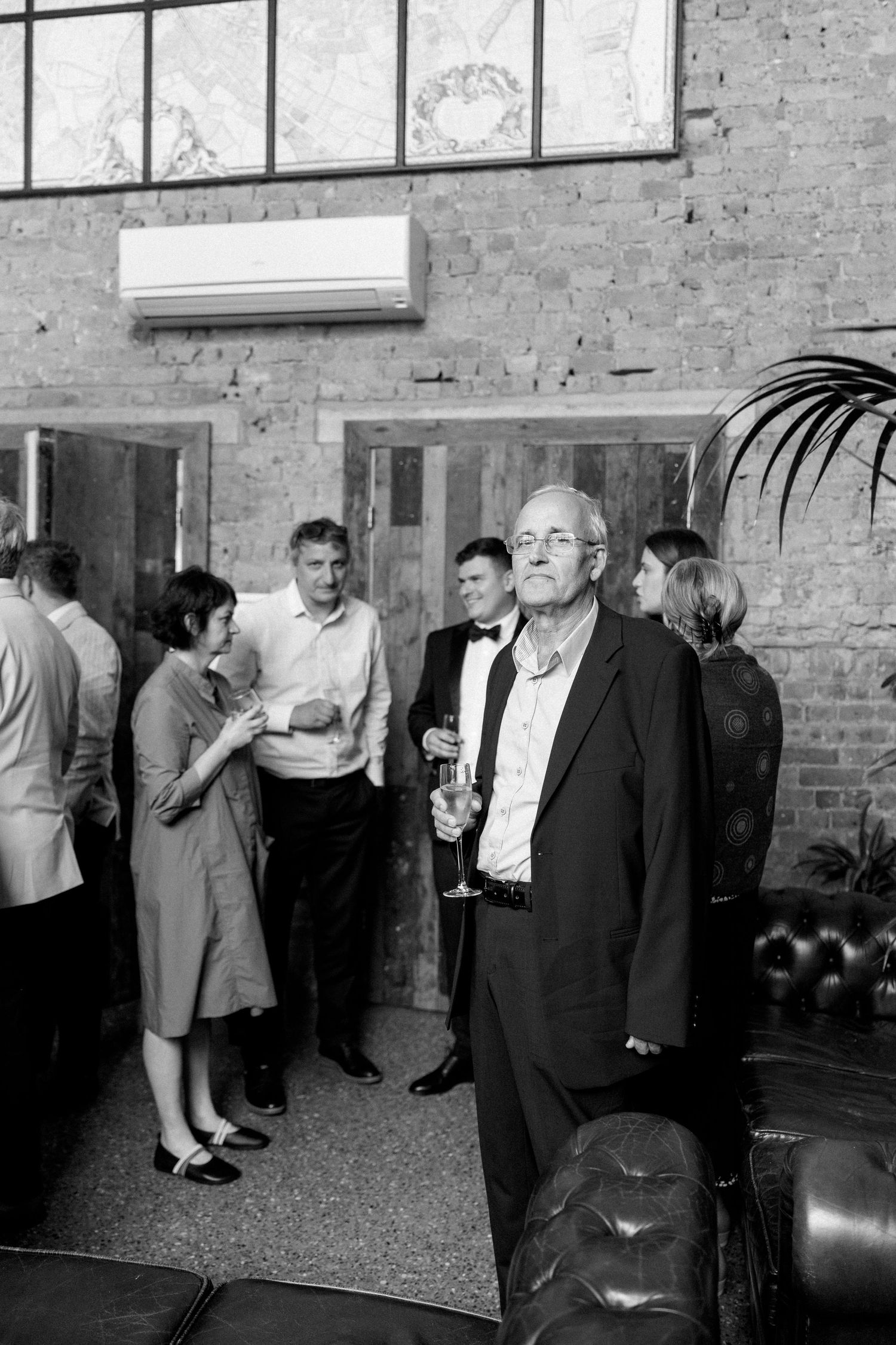 Black and white photo of guests mingling at an indoor reception with brick walls.
