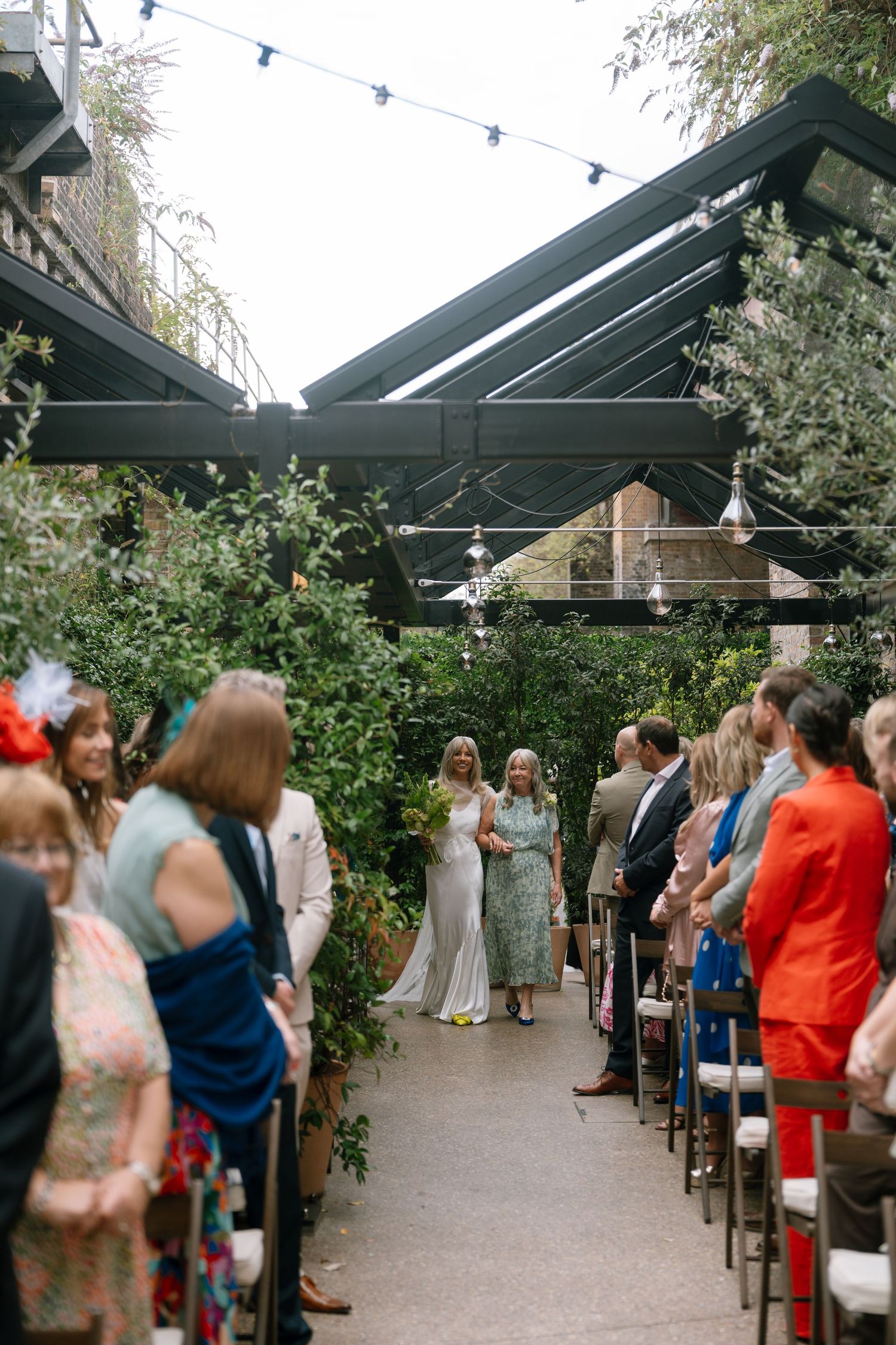 Outdoor wedding ceremony setup with guests seated under a covered pergola with string lights.