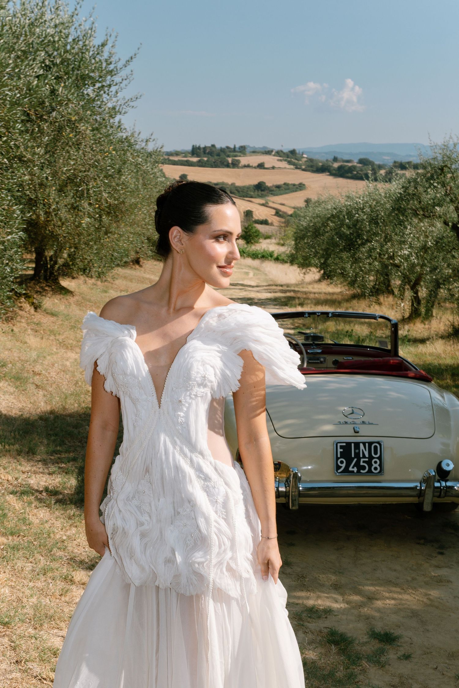 Classic vintage car parked in a sun-drenched olive grove with rolling Tuscan hills in the background.