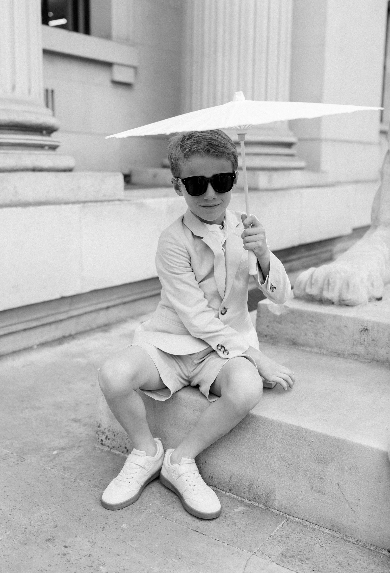 A stylish figure in sunglasses holds a white parasol while sitting on stone steps.