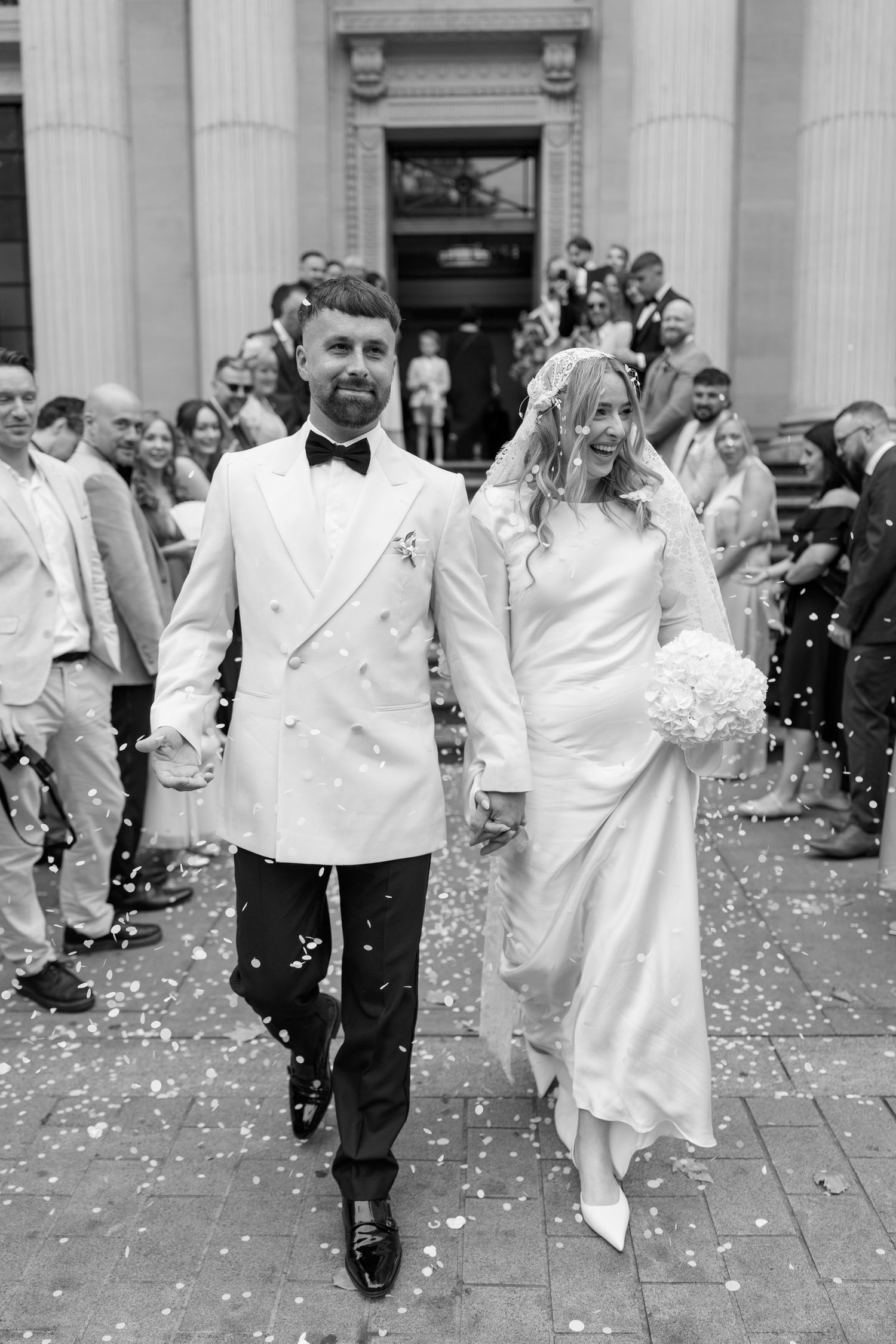 Newlyweds exiting building while guests throw white confetti on stone steps.