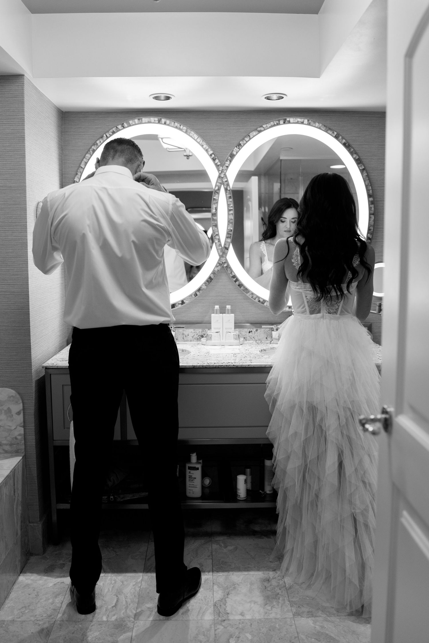 A couple getting ready in a bathroom with two circular mirrors, the bride wearing a flowing white dress.