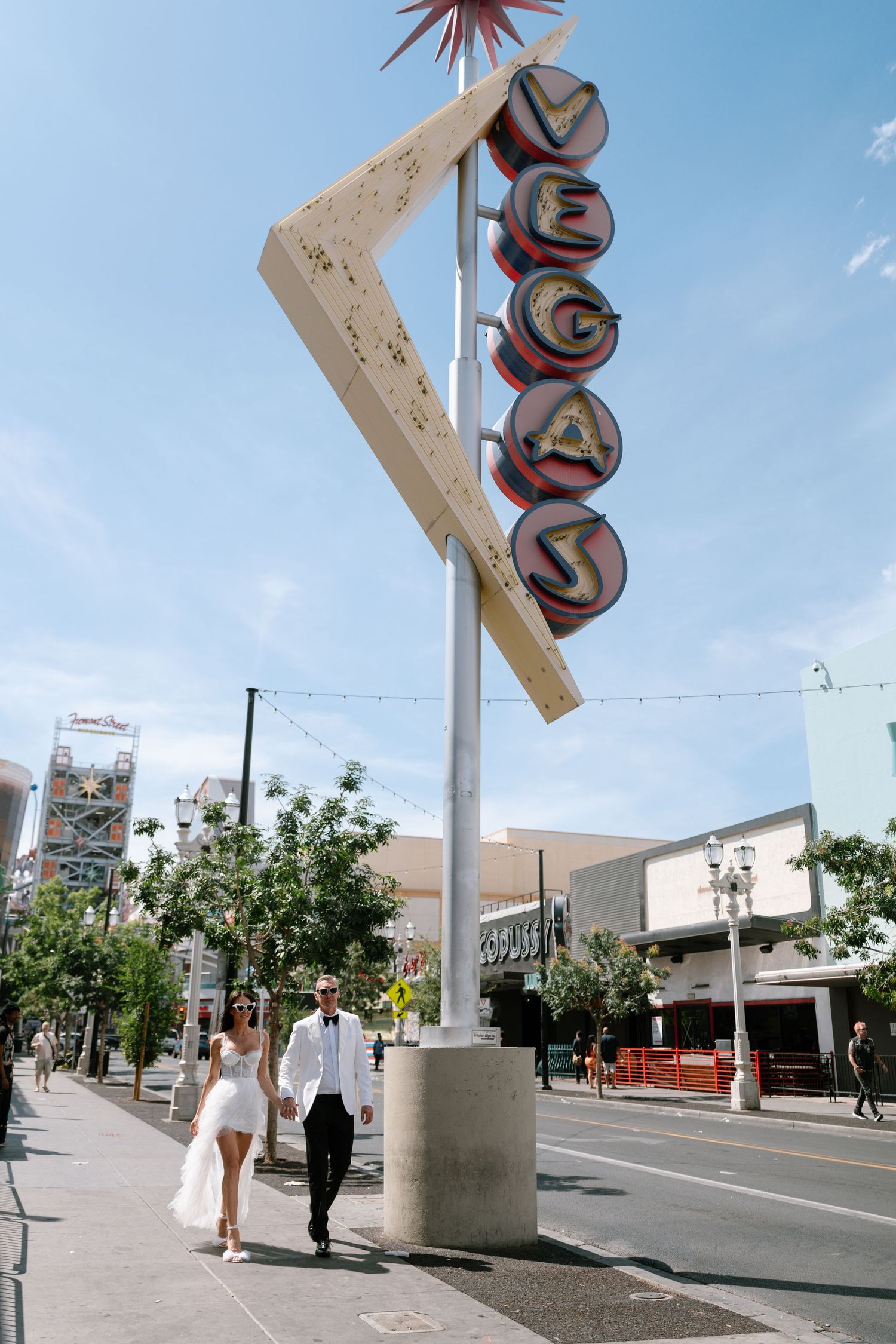 Couple in wedding attire walking beneath retro Vegas sign on sunny street.